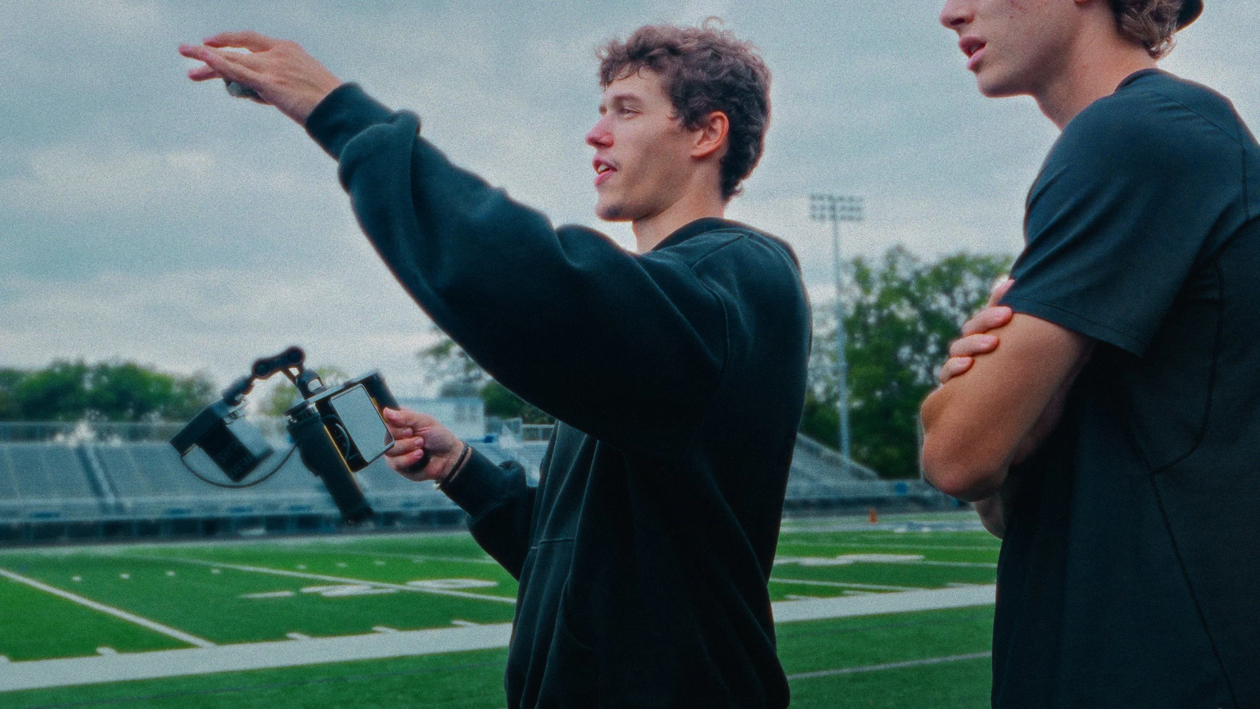 Two young men on a sports field; one is holding a directors monitor and showing the other a mobile device, with cloudy sky overhead.