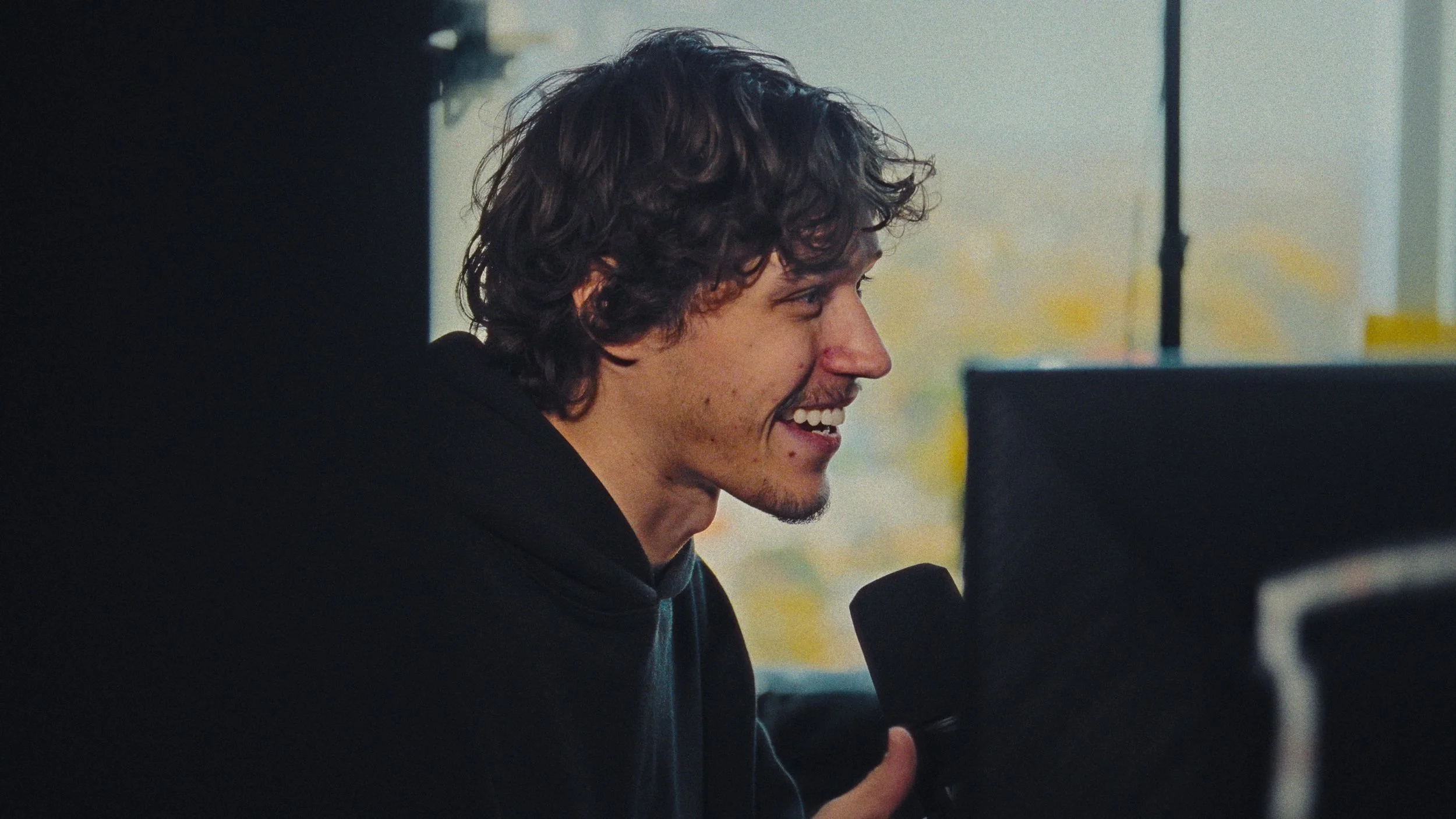 A young man with curly brown hair smiling and holding a microphone in front of a computer monitor.