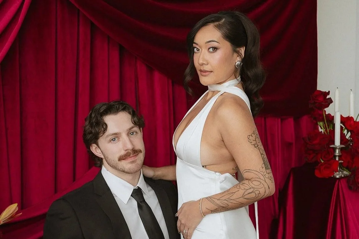 A woman in a white wedding dress with tattoos on her arm and shoulder, standing next to a man in a black suit and white shirt, against a red velvet curtain with a lit candelabrum and red roses nearby.