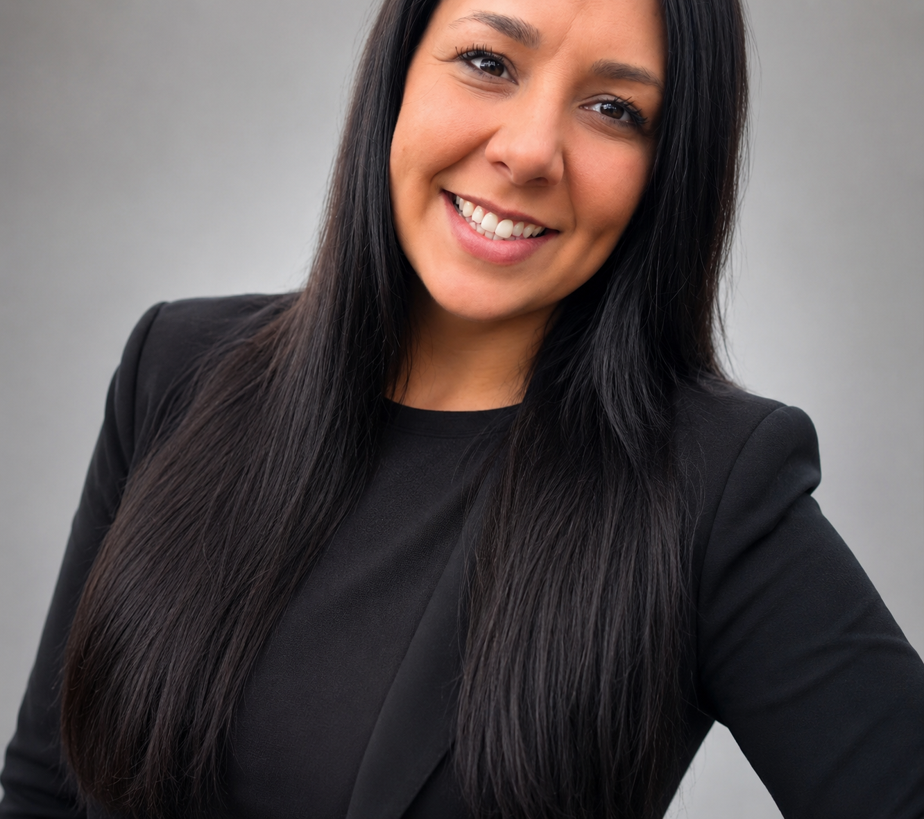 Close-up portrait of a smiling woman with long black hair wearing a black blazer and top, standing against a gray background.