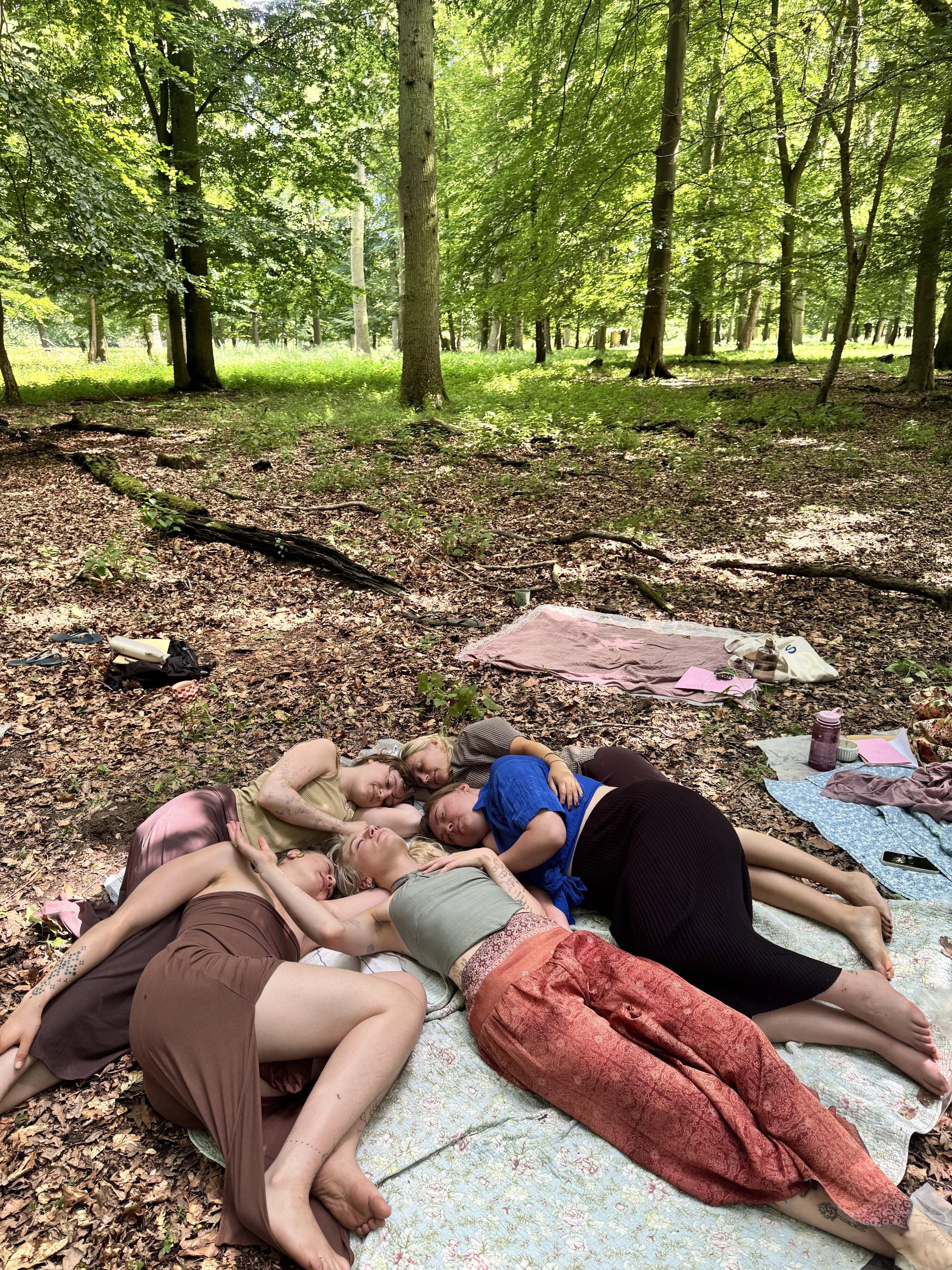 Five young women lying closely together on blankets spread on the forest floor, with trees and sunlight in the background.