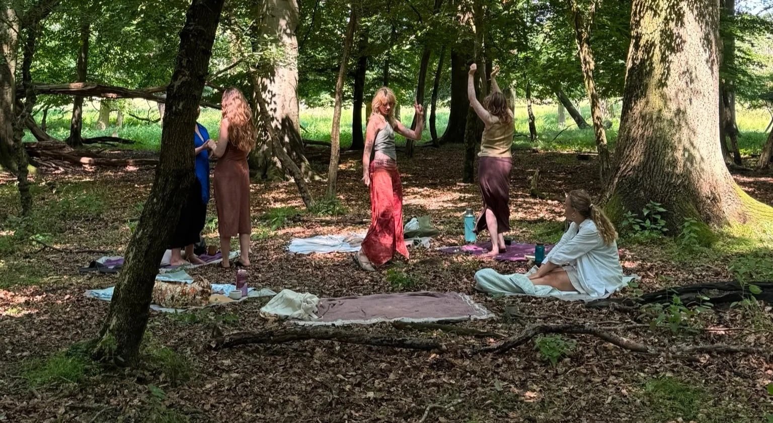 Group of women in a circle in a forest with tall trees and green leaves, some standing and some sitting on mats.
