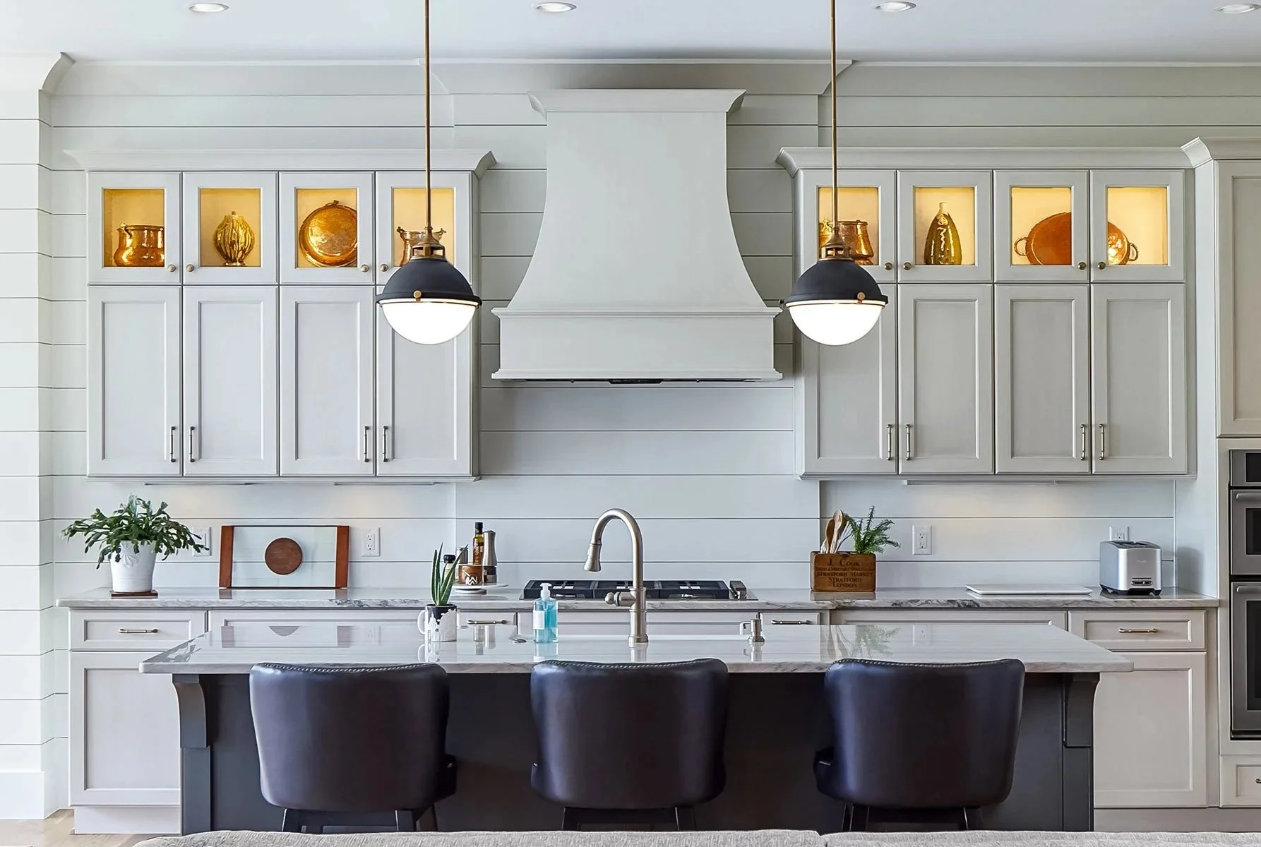 Modern white kitchen with black pendant lights, decorative gold plates in glass cabinets, and a kitchen island with black chairs.