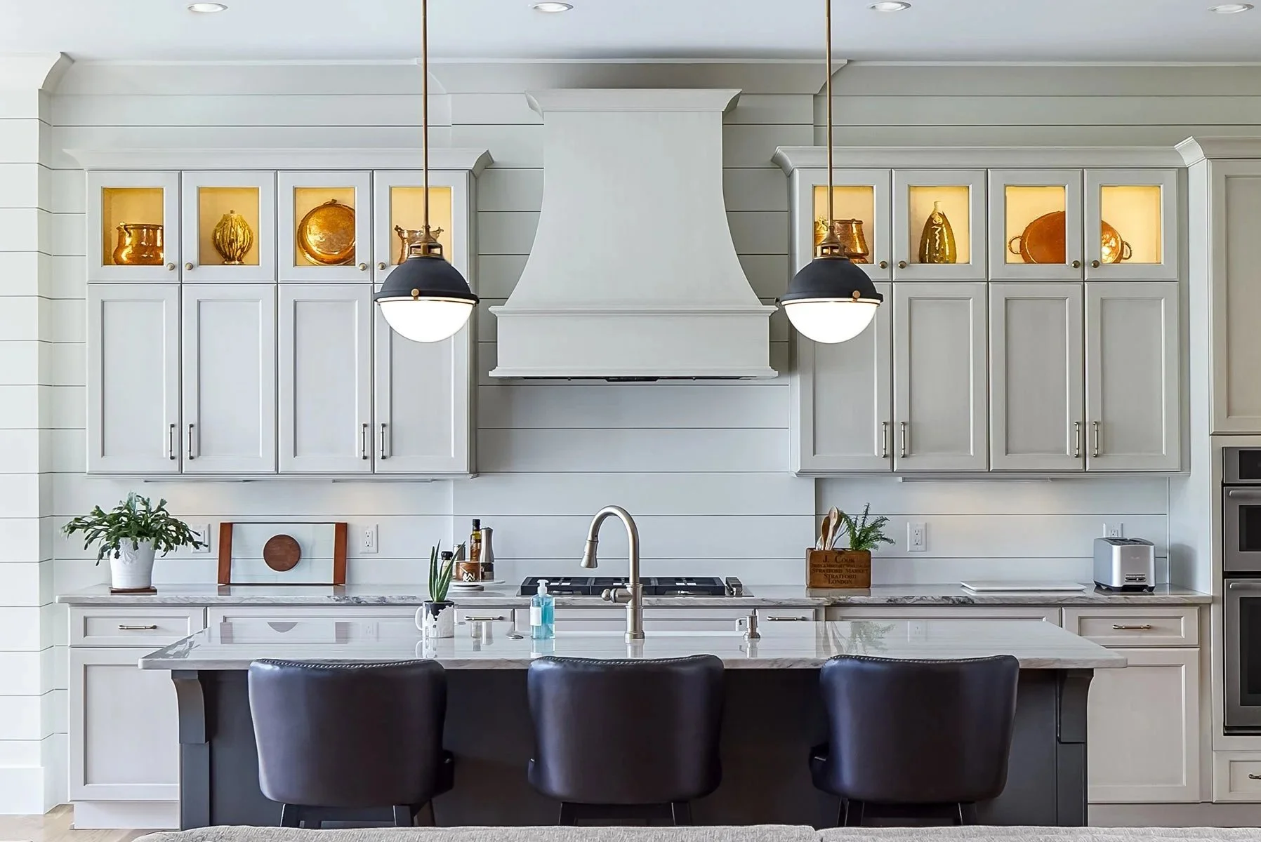 Modern white kitchen with high cabinets featuring glass-fronted sections displaying gold-colored decorative items, a central white range hood, two black pendant lights, marble countertops with plants and kitchen essentials, a stainless steel toaster, and three black chairs at a kitchen island.
