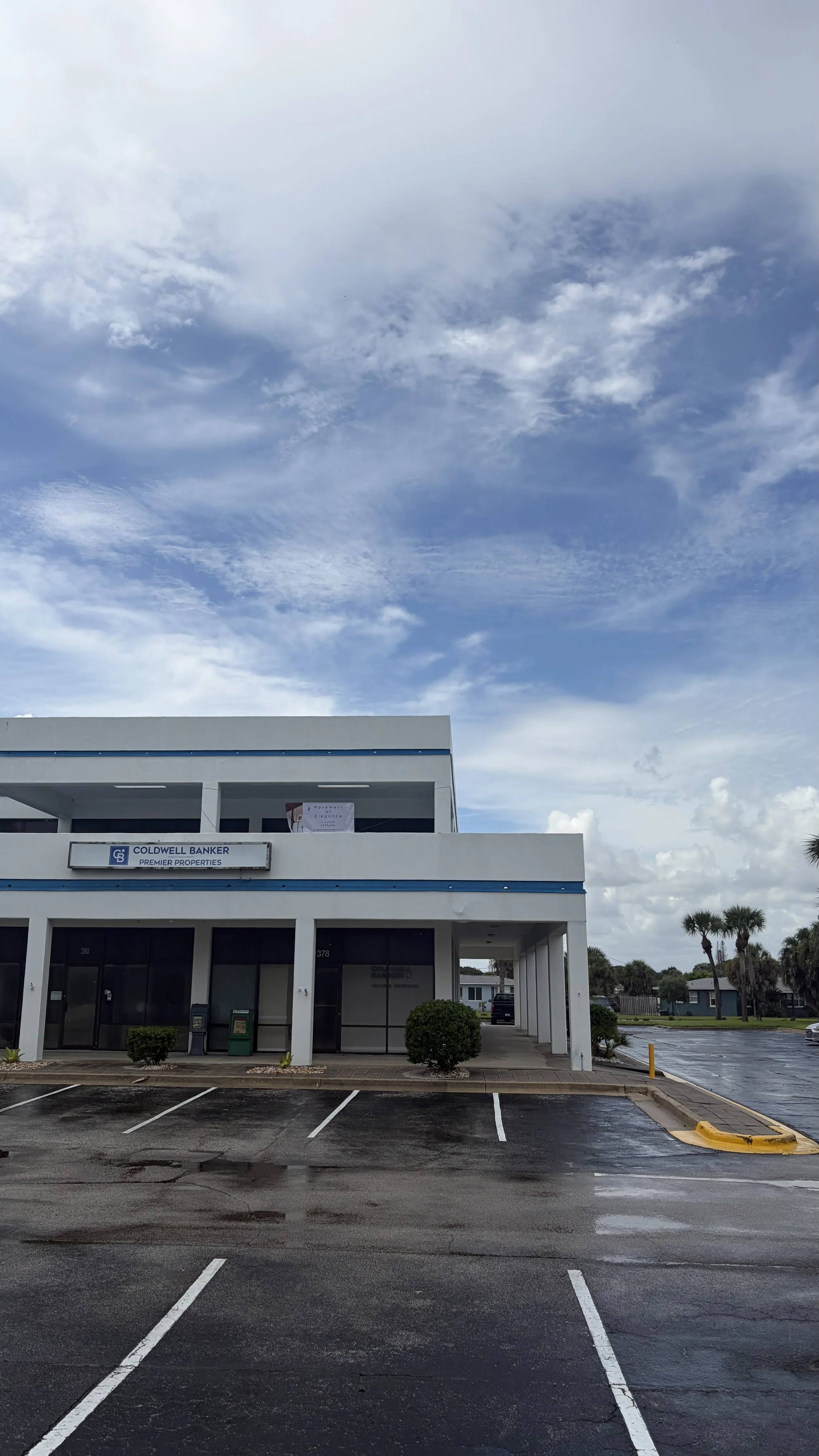 Empty parking lot in front of a two-story commercial building with a sign that reads Coldwell Banker Premier Properties. The sky is partly cloudy with a mostly blue sky and scattered clouds.