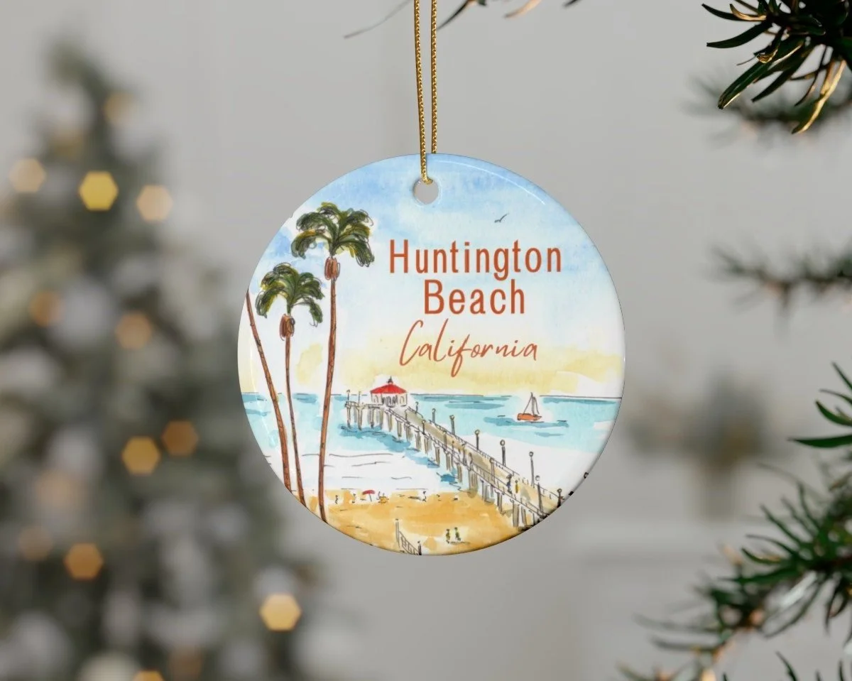 A circular ornament with a watercolor illustration of Huntington Beach, California, showing palm trees, a pier, and a boat on the water, hanging from a Christmas tree.