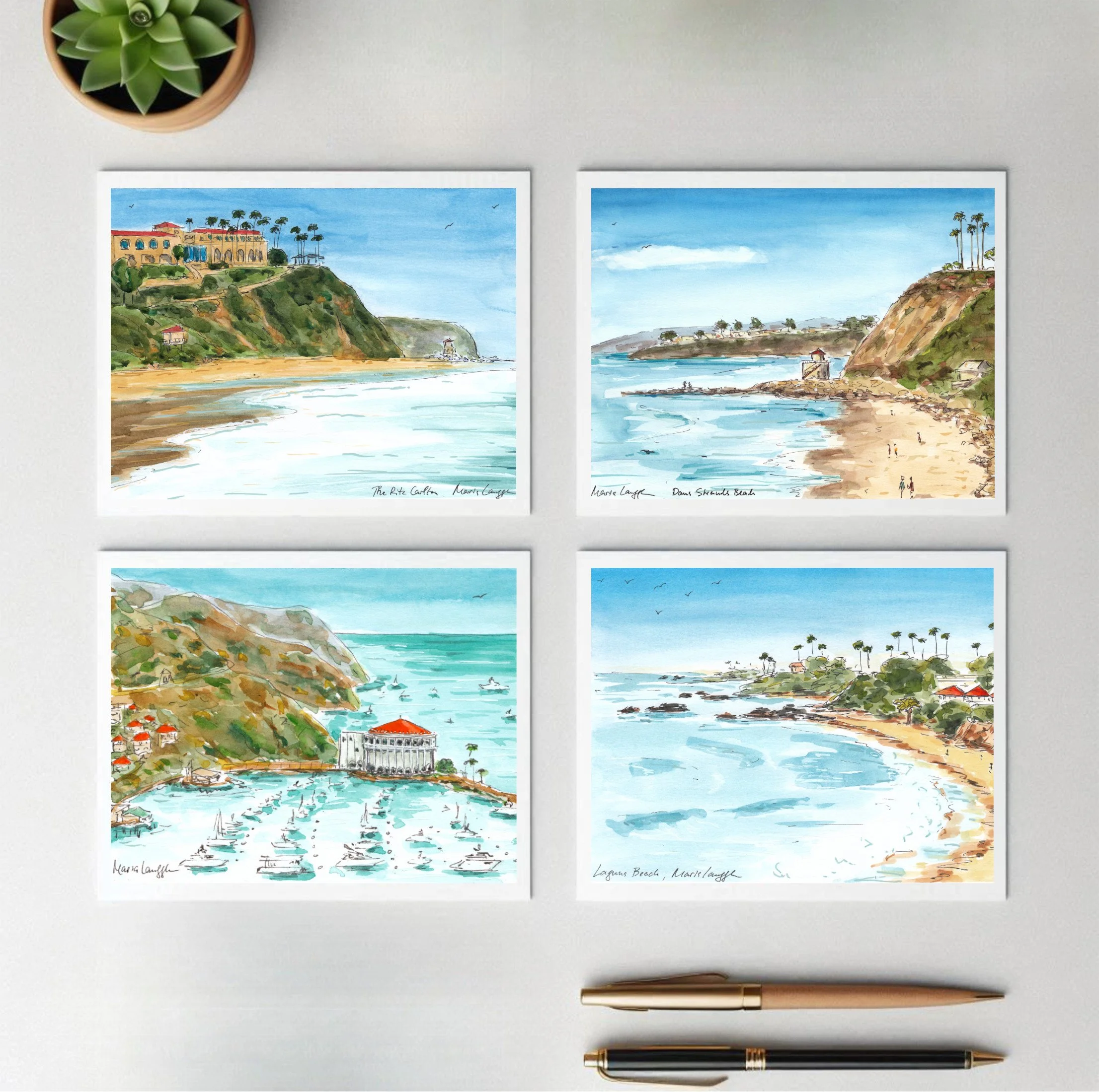 Four watercolor landscape paintings of coastal scenes with beaches, cliffs, and ocean views, labeled with locations like Malibu, Laguna Beach, and Dana Strands Beach, arranged on a white wall with a potted succulent plant above and pens below.