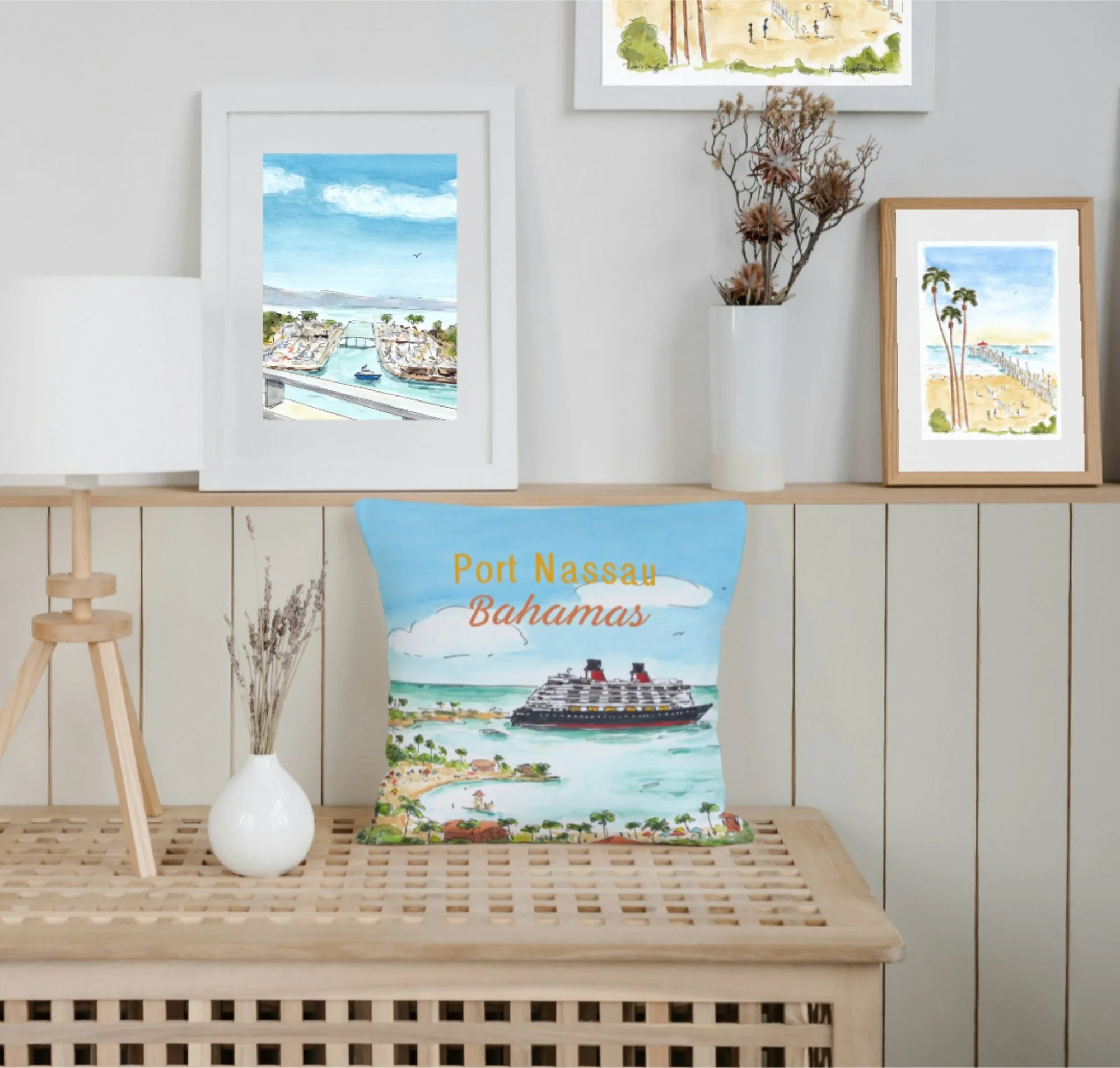Decorative room with framed coastal artwork, a white vase with dried flowers, a lamp, and a pillow with a Port Nassau Bahamas cruise ship illustration on a wooden table.