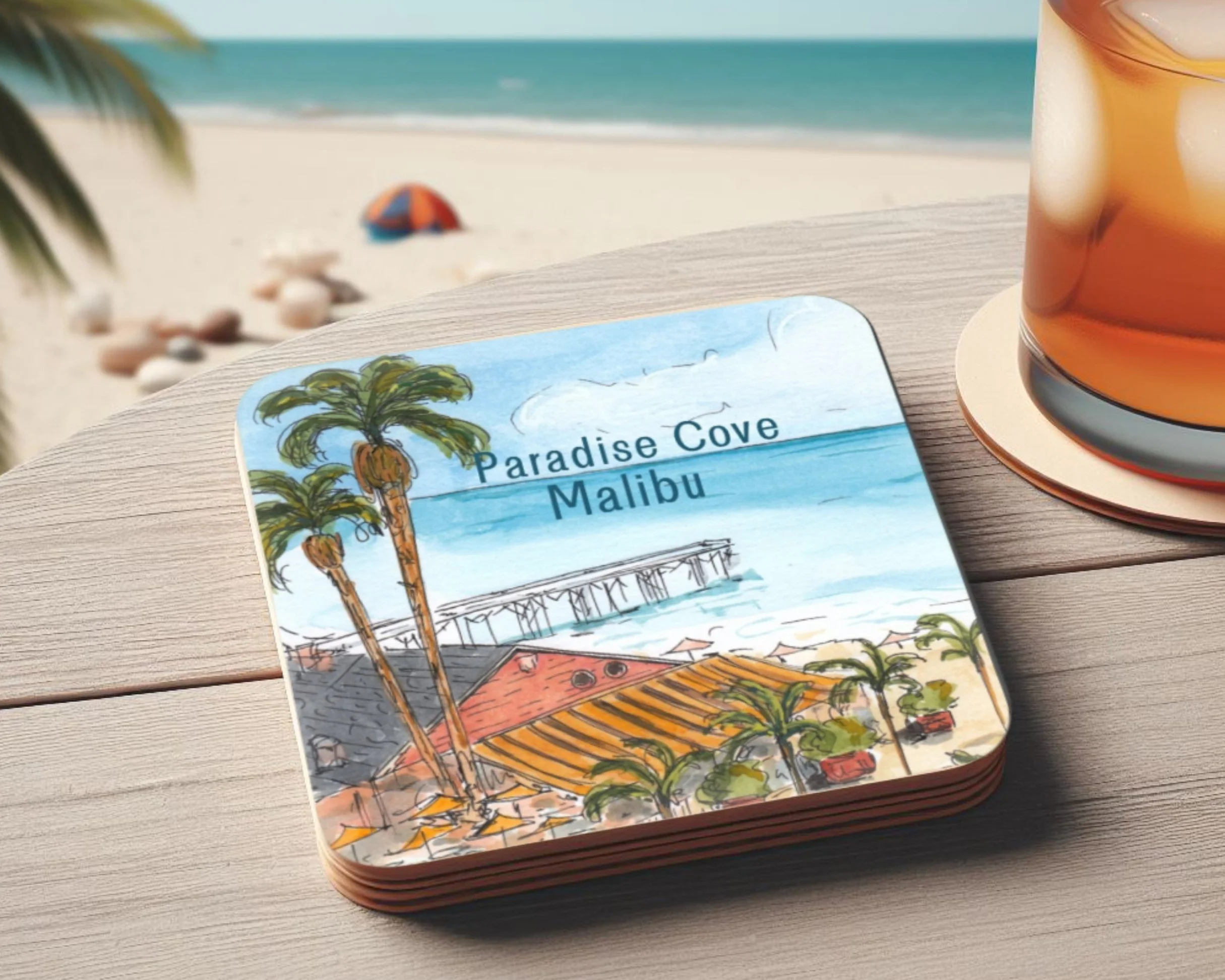Illustrated coaster with palm trees, a pier, and beach scene labeled 'Paradise Cove Malibu', placed on a wooden table next to a glass of iced tea, with a beach and umbrella in the background.