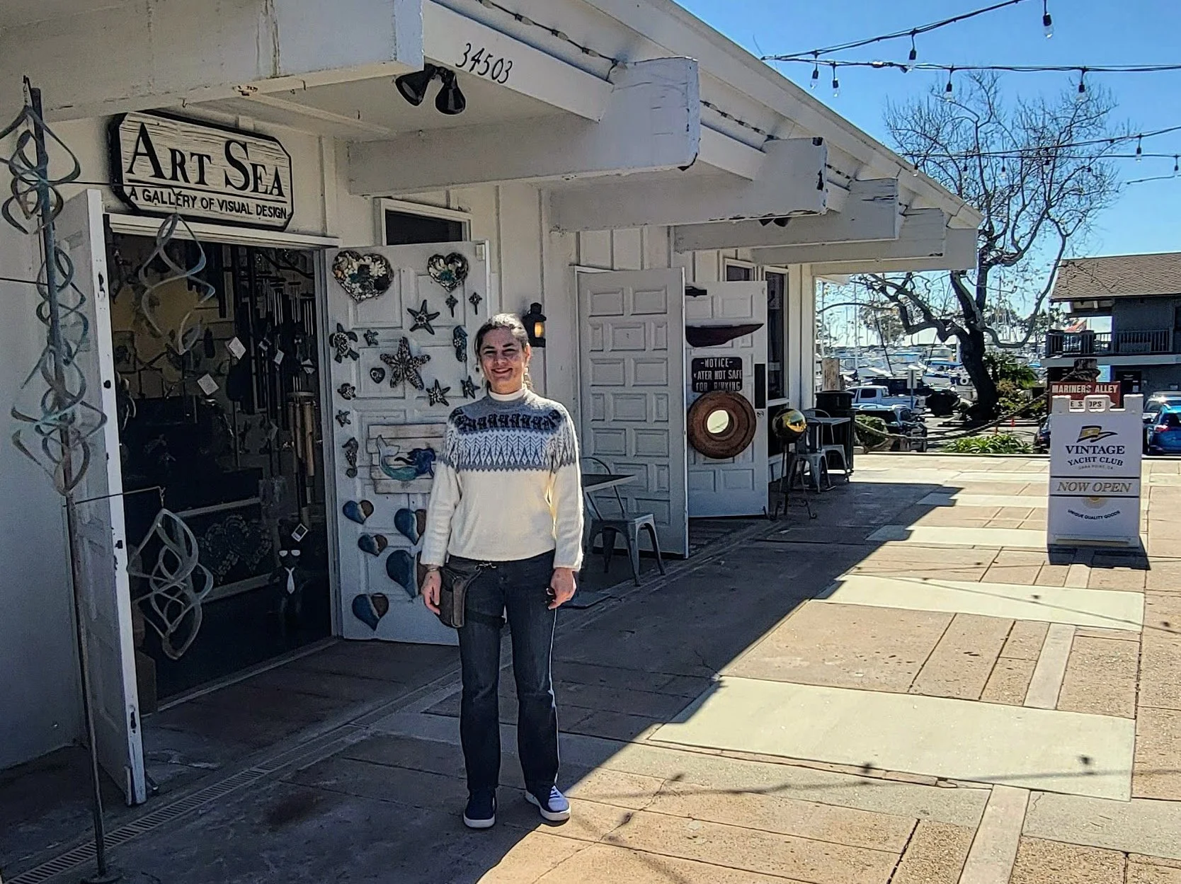 A woman standing in front of the Art Sea gallery, smiling. The gallery has decorations on the door and wall, with a sign indicating it's open. The scene is outdoors with a clear sky, nearby trees, and parked cars.