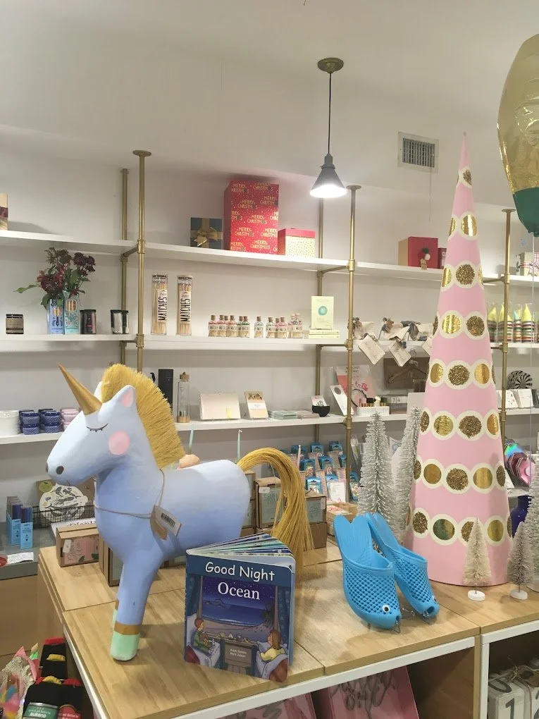 Display of unicorn-themed and holiday decorations in a store, including a unicorn-shaped light, a pink and gold Christmas tree, and blue whale-shaped slippers.