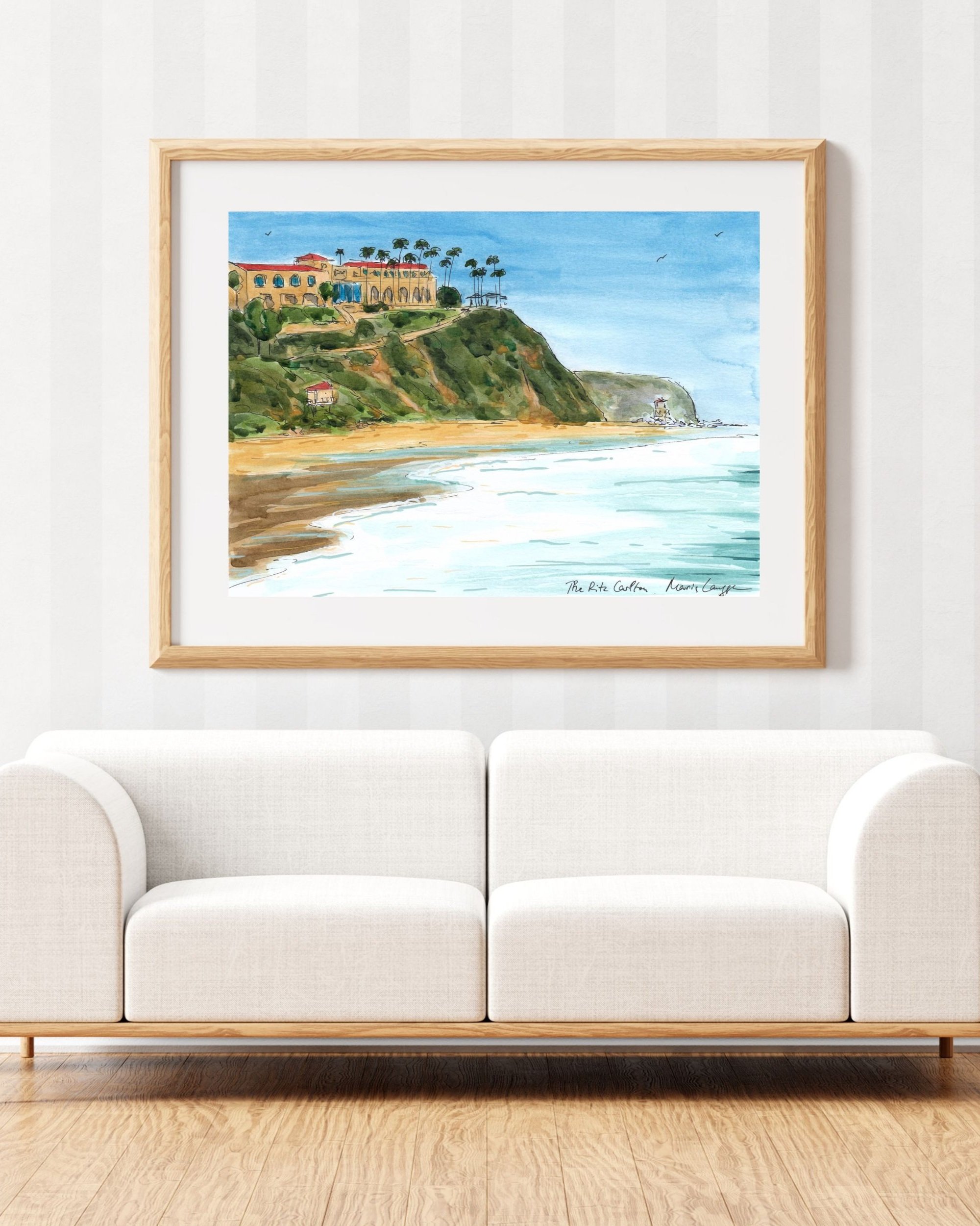 A framed watercolor painting of a beach with cliffs, trees, and buildings hanging above a white sofa.