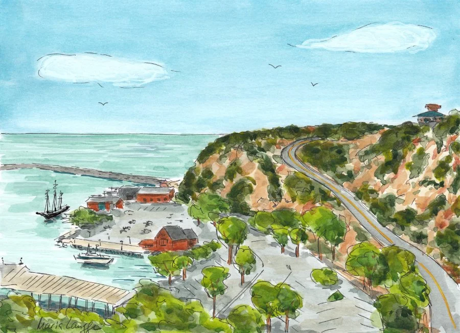Waterfront scene with boats docked near red buildings, a hilly coastline with trees, and a winding road along the hills, under a blue sky with a few clouds and birds flying.