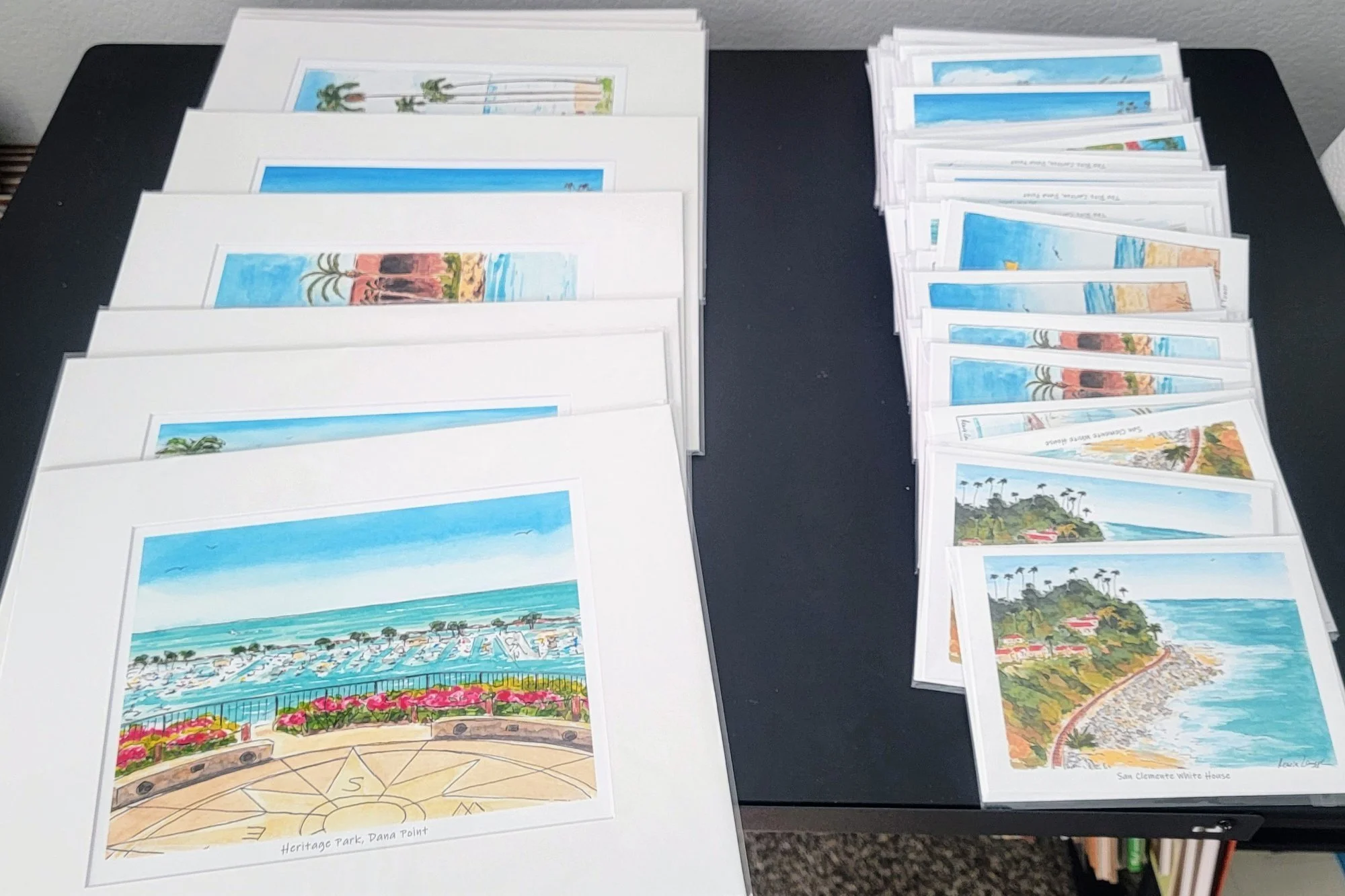 Printouts of landscape watercolor illustrations of coastal scenes, a beach with umbrellas, and a park with a compass rose, arranged on a black table.