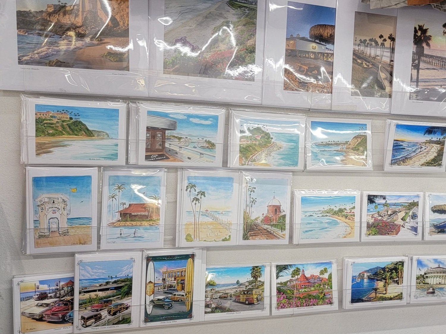 Collection of landscape paintings and photographs depicting coastal scenes, beach views, palm trees, and iconic California landmarks, displayed in clear plastic sleeves on a wall.