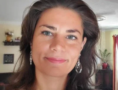 A woman with long brown hair and blue eyes, smiling, wearing silver earrings, standing in a home interior with shelves and a plant in the background.