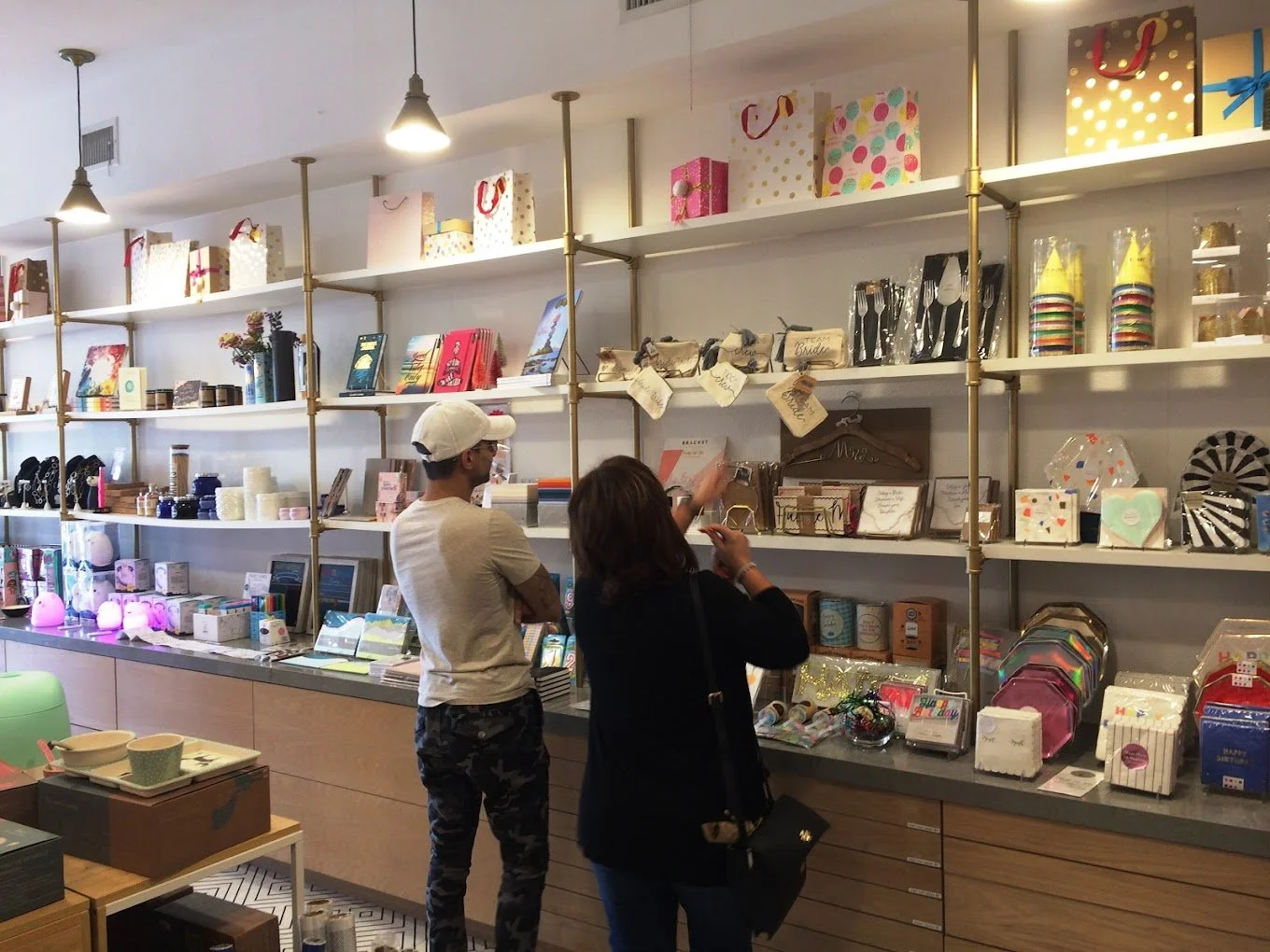 Two people shopping at a store with shelves filled with gifts, home decor, and party supplies. Items include gift bags, books, candles, kitchenware, and colorful party hats.