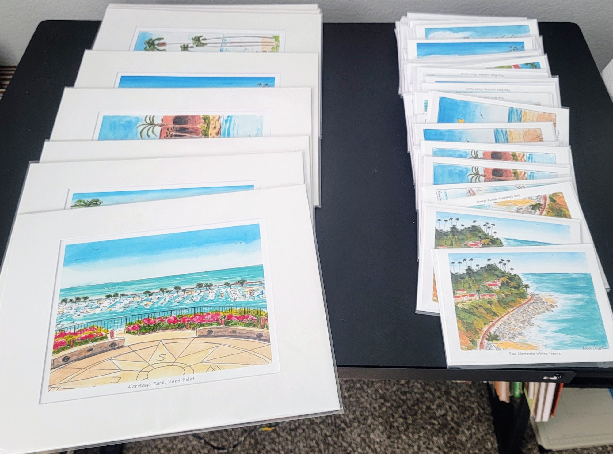 Collection of framed watercolor paintings of coastal landscapes and beaches, organized on a black table.