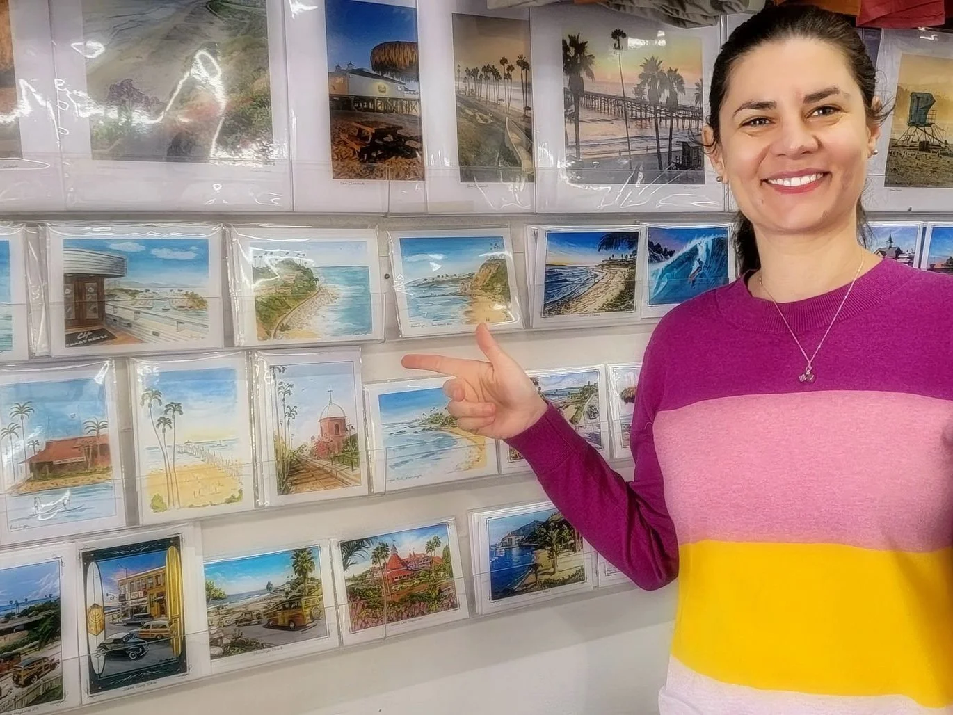 A woman smiling and pointing at a display of various scenic postcards on a wall.
