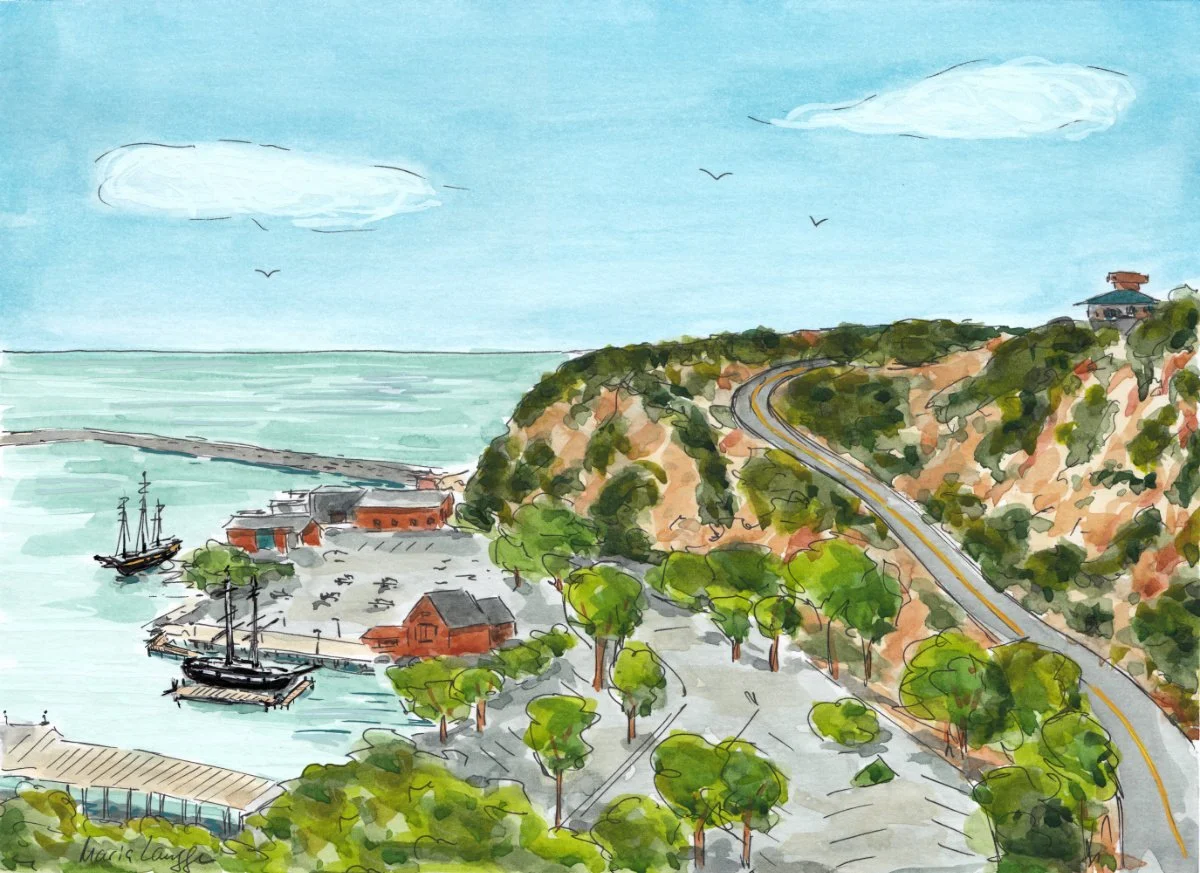 Waterfront scene with boats docked, a parking lot, trees, a winding road on a hillside, and a building on top of the hill under a partly cloudy sky.
