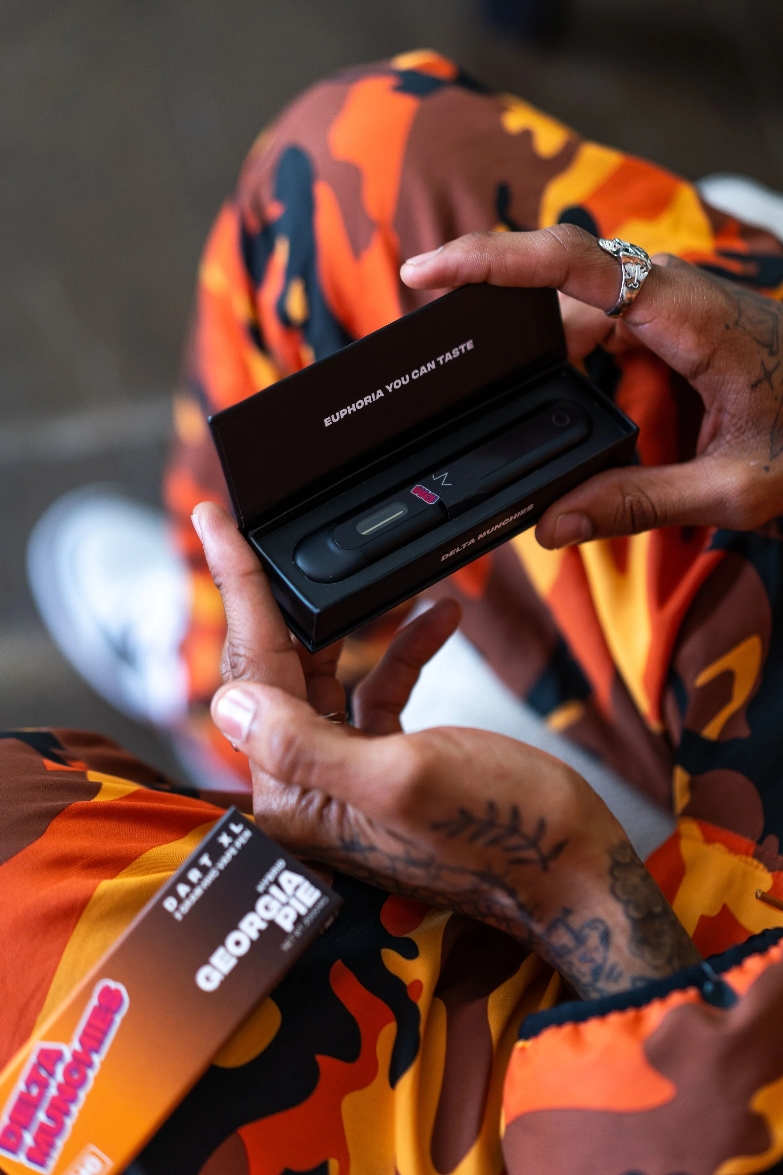 A person holding a black vape pen in an open box labeled 'Euphoria You Can Taste,' with decorative tattoos visible on their hand. They are wearing orange camo pants. The box mentions 'Delta Munchies' and 'Georgia Pie.'"