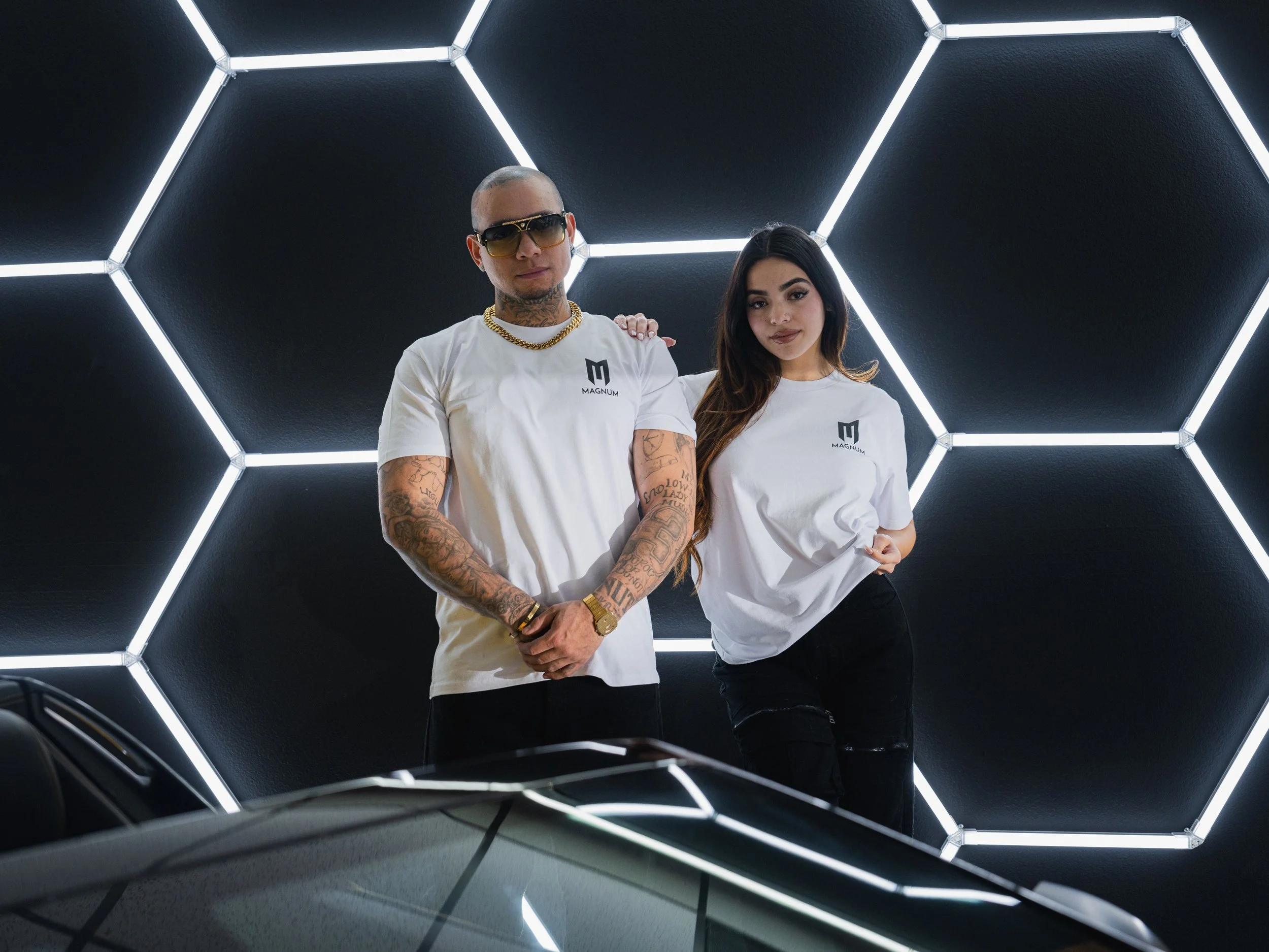 A man and a woman posing in front of a hexagonal LED light wall, both wearing white Magnum T-shirts. The man has tattoos and sunglasses, and both appear to be in a car showroom. The car roof is partially visible.