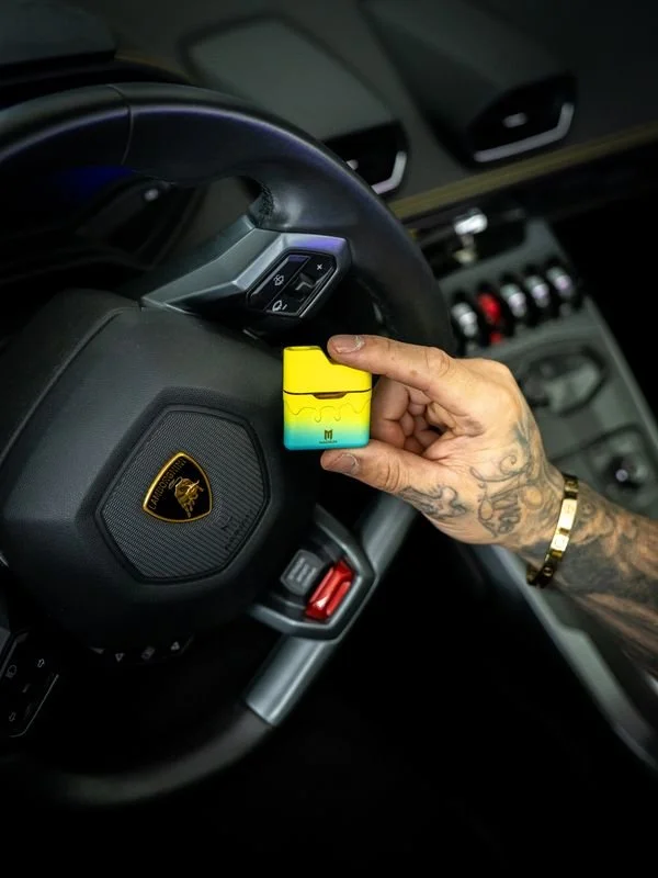 Tattooed hand holding a yellow and blue vape pod inside a luxury car with a Lamborghini steering wheel.