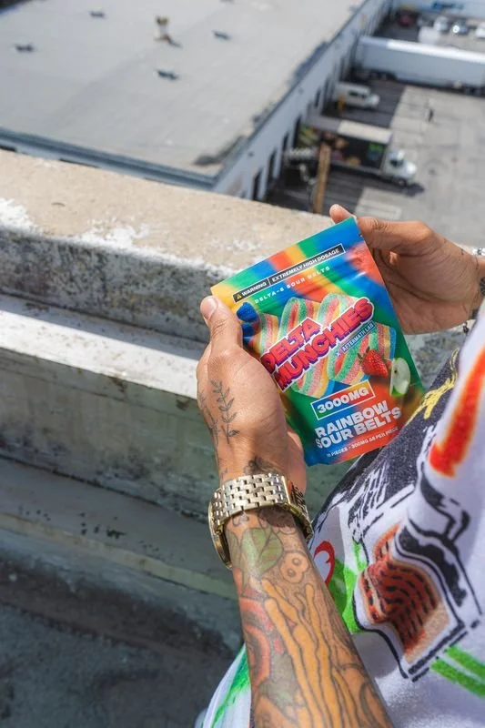 Person holding a colorful package of Delta Munchies Rainbow Sour Belts on a rooftop.