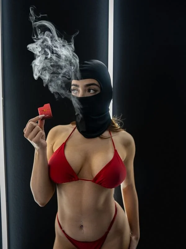 Person wearing red bikini and black balaclava, holding a red cigarette box with smoke.