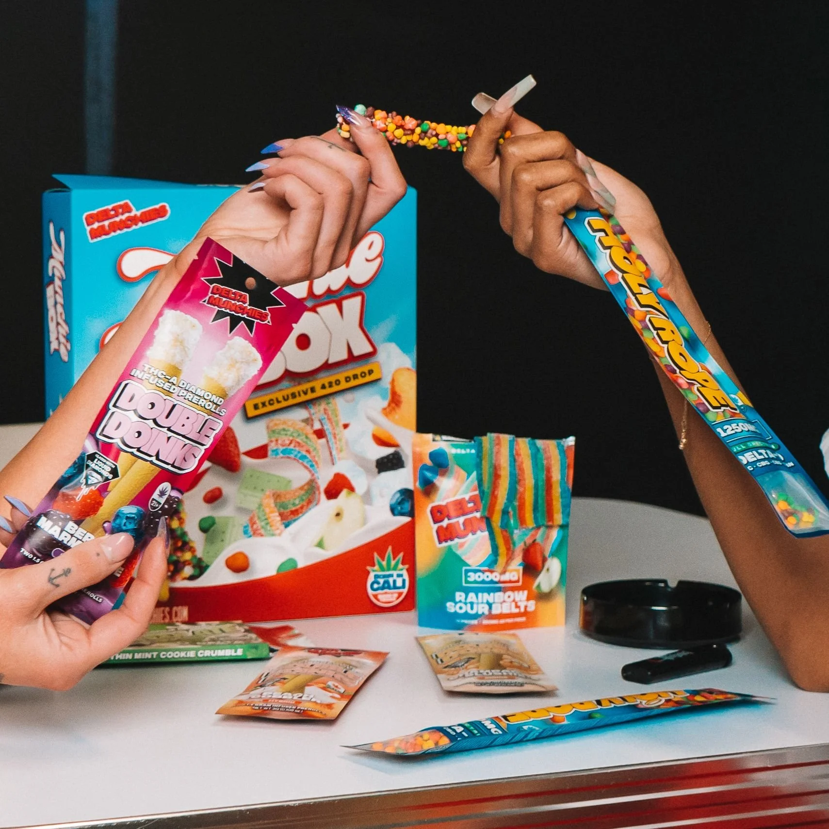 Hands holding colorful candy, including packages labeled "Double Donks" and "Holy Rope," alongside a candy assortment box and packaging for "Rainbow Sour Belts" on a table.