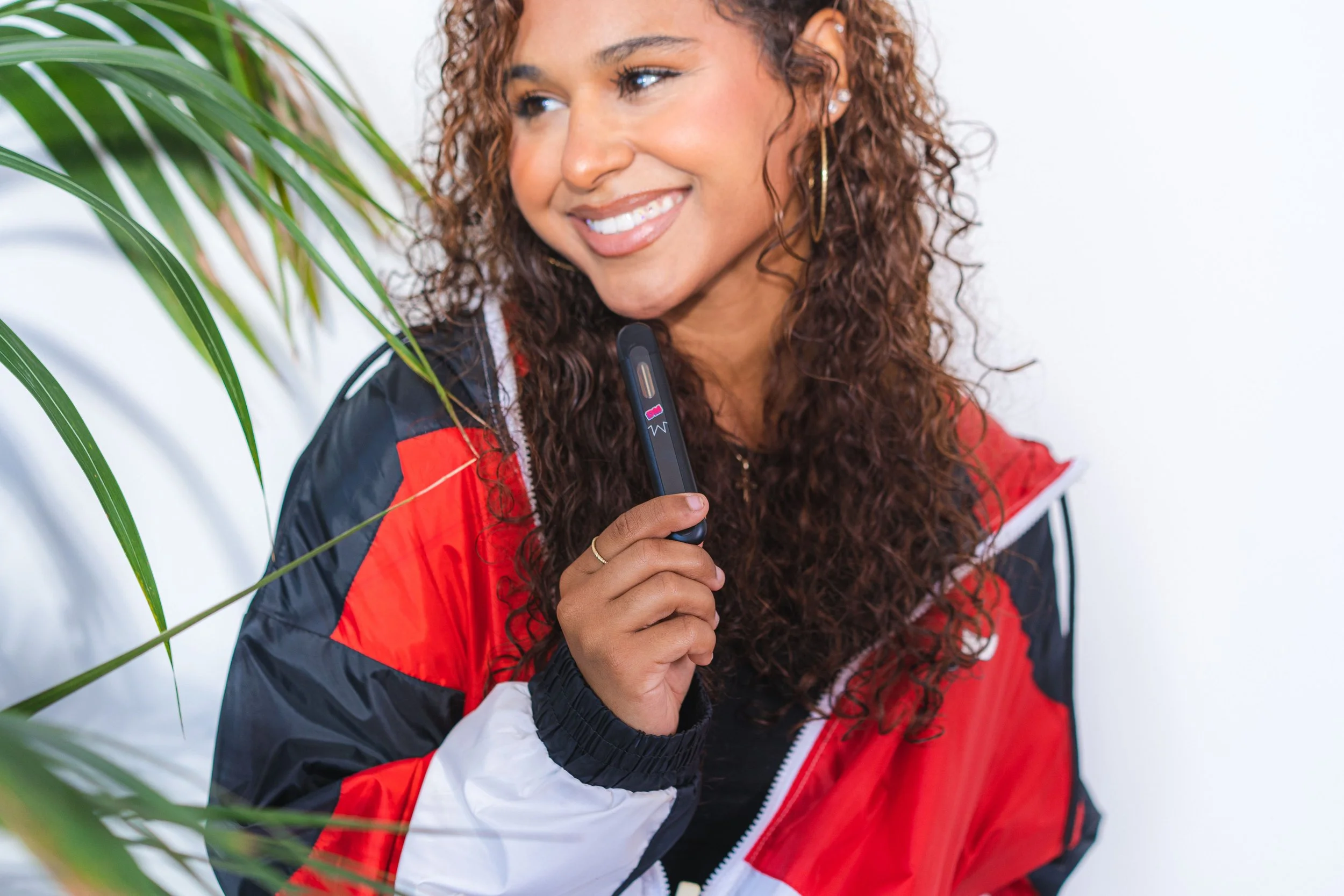 Woman smiling and holding a vape pen, wearing a red, black, and white jacket, with a leafy plant in the foreground.