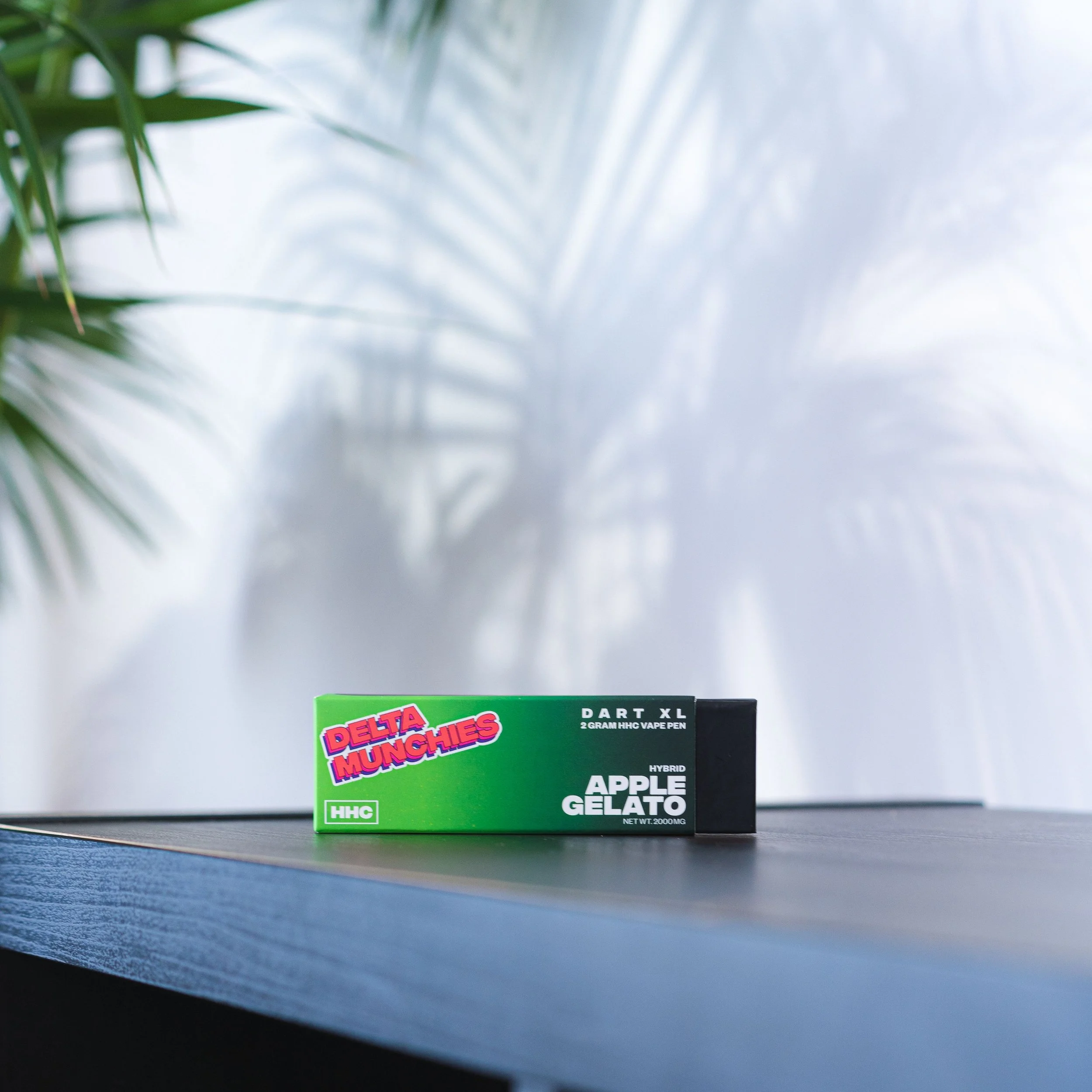 Box of Delta Munchies Apple Gelato HHC vape pen, placed on a dark wooden surface with plant shadows on the wall.