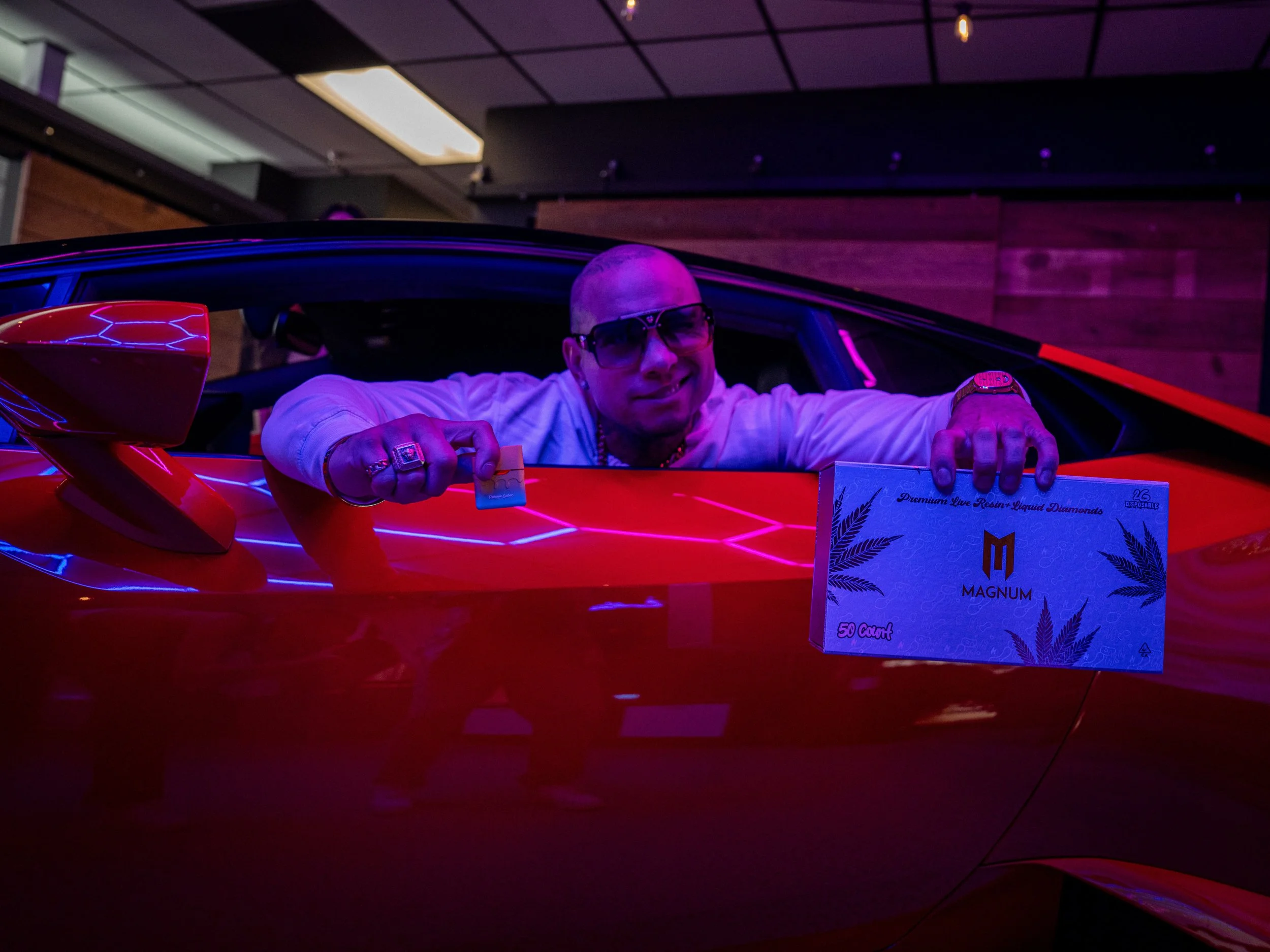 Person leaning out of a car window holding a Magnum package and a card, under neon lighting.