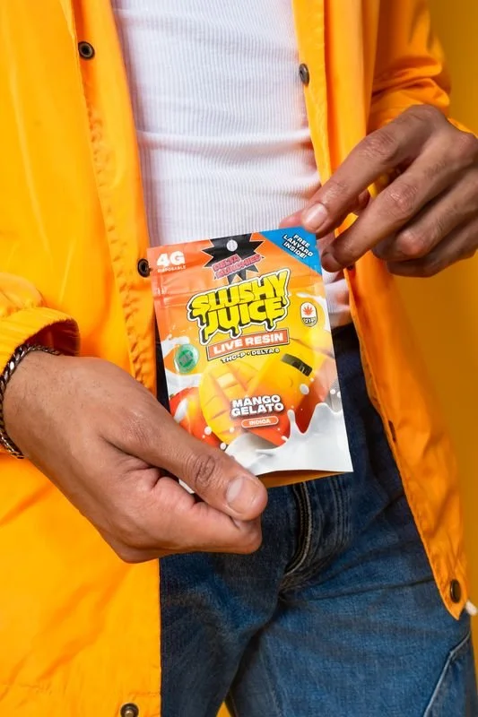 Person holding a package of Slushy Juice live resin vape cartridge flavored with Mango Gelato, wearing an orange jacket and jeans.