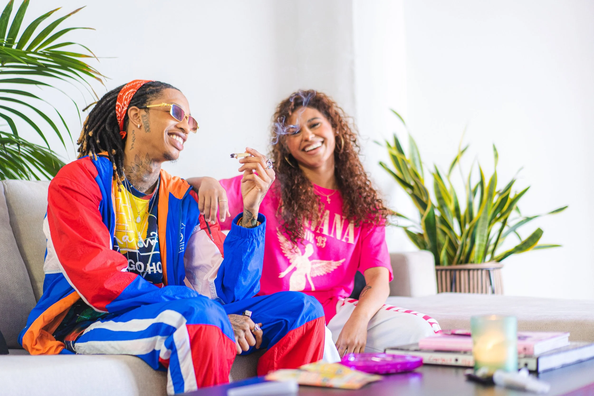 Two individuals sitting on a sofa, smiling and holding a smoking item, with indoor plants and books on a table nearby.