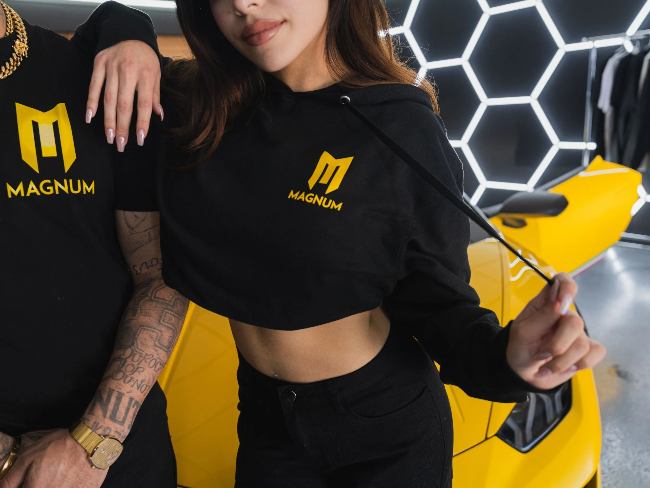 Two people wearing black "Magnum" branded shirts stand in front of a yellow sports car with a honeycomb-patterned light background.