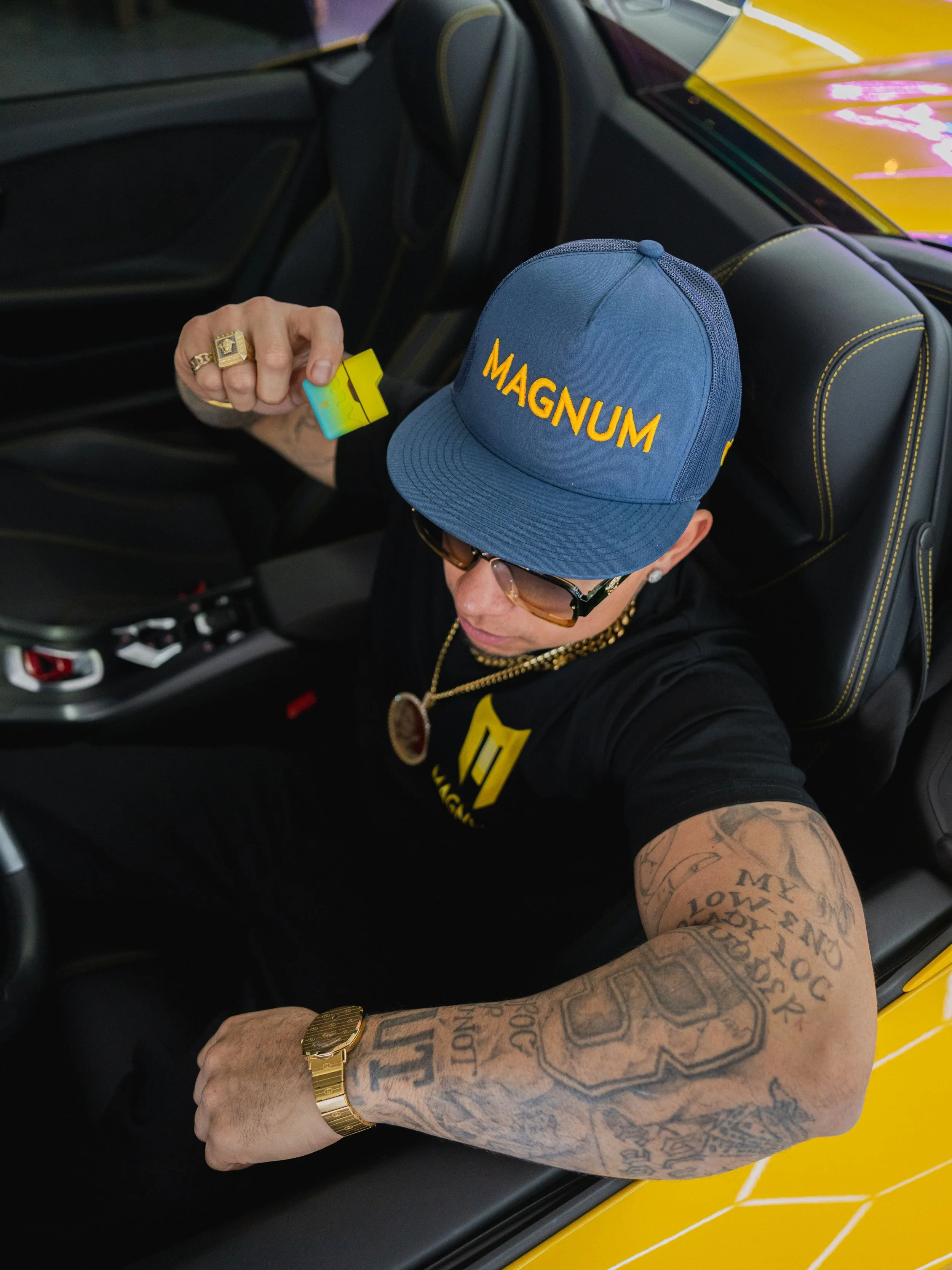 A person sitting in a yellow car, wearing a blue cap with 'MAGNUM' written on it, sunglasses, and a black T-shirt with a logo. The person has tattoos on the right arm and is holding a small yellow and blue card. The person is also wearing gold jewelr