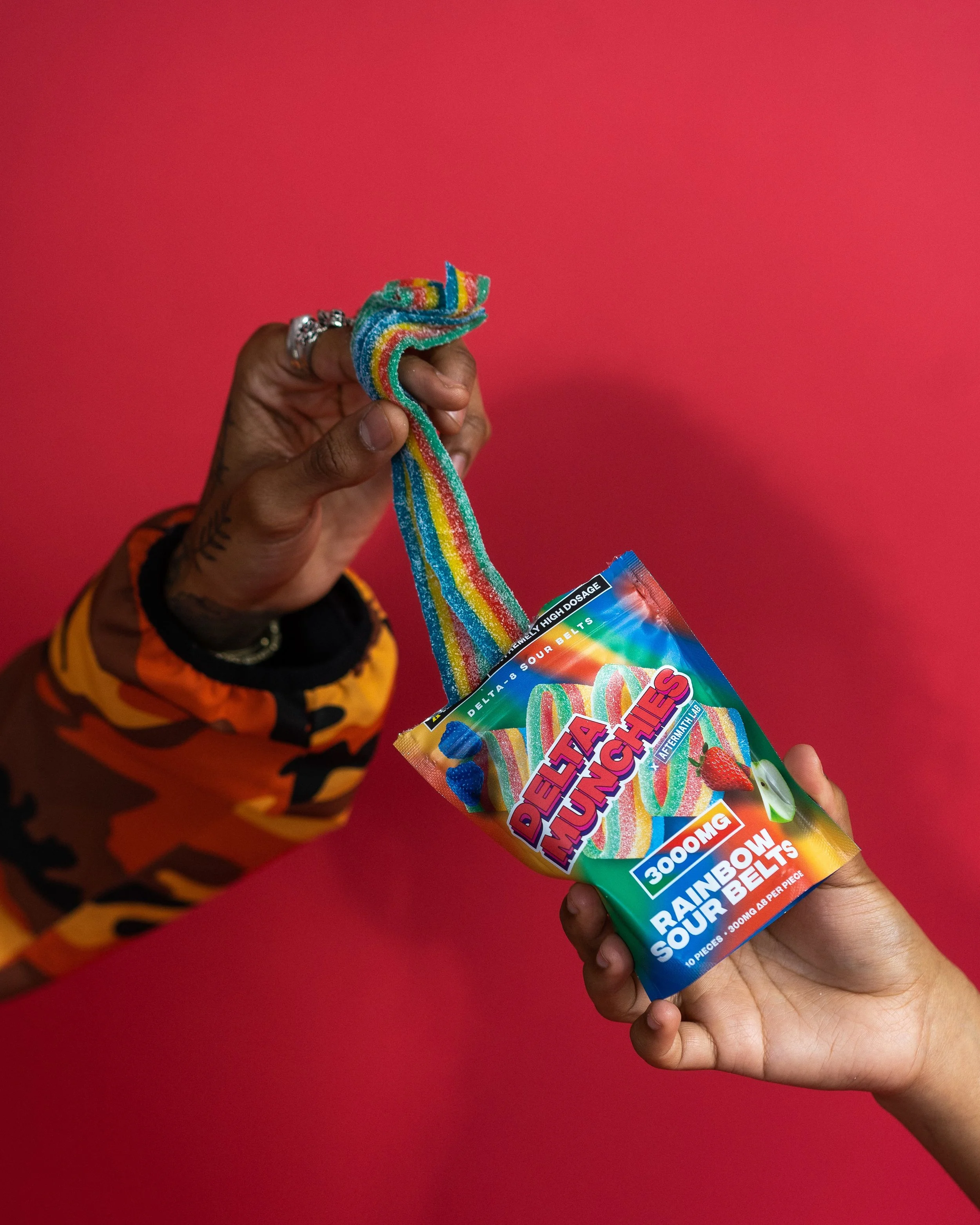 Two hands holding Delta Munchies Rainbow Sour Belts package against red background.