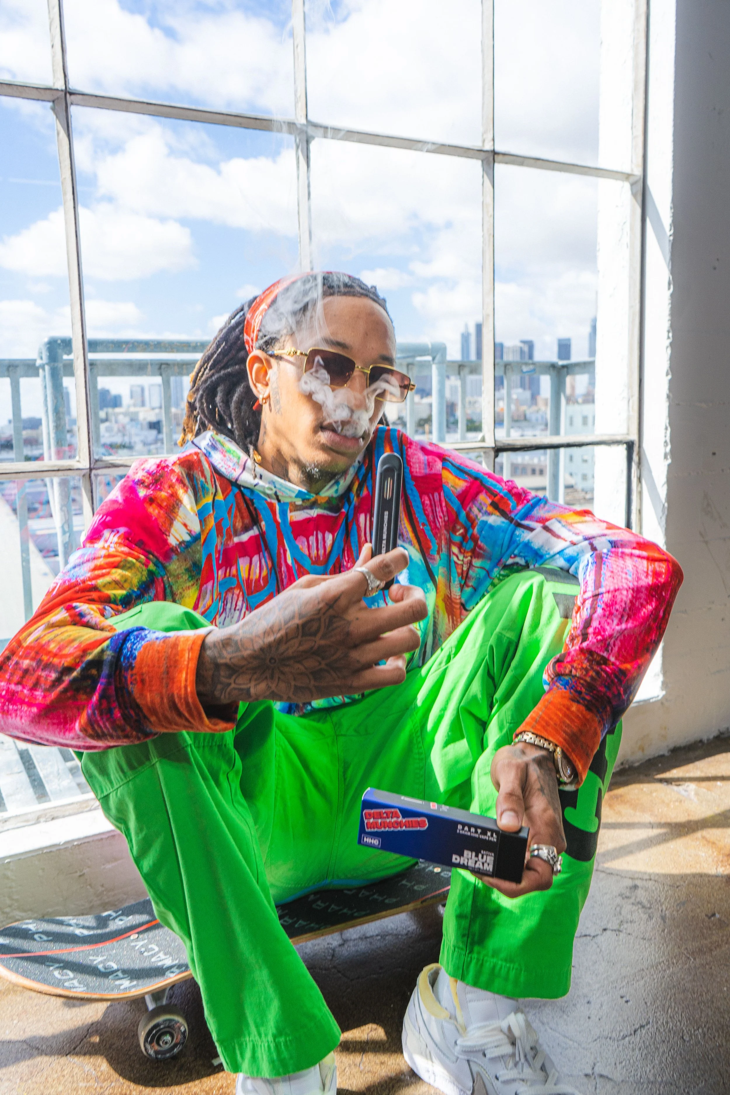 Person sitting on a skateboard indoors, wearing colorful clothing and sunglasses, holding a vape pen, with smoke around their face.