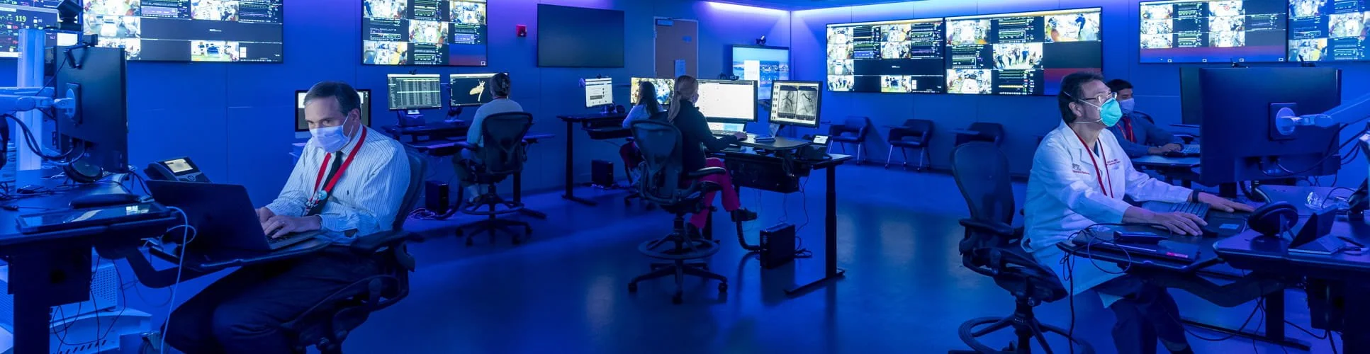 A technician wearing an N95 mask sits at a desk and monitors a screen in the CICU telehealth command center.