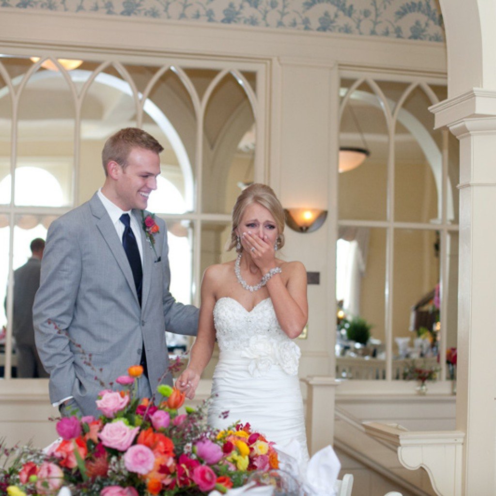 A bride and groom standing together indoors. The bride is wearing a white strapless wedding dress and jewelry, covering her mouth with her hand, looking emotional. The groom is in a light gray suit with a boutonniere, smiling. There is a colorful flo
