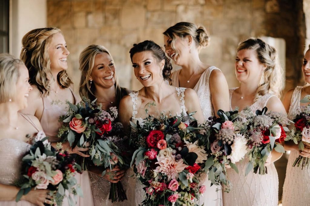 Group of bridesmaids and bride smiling together, holding bouquets, wearing light-colored dresses.