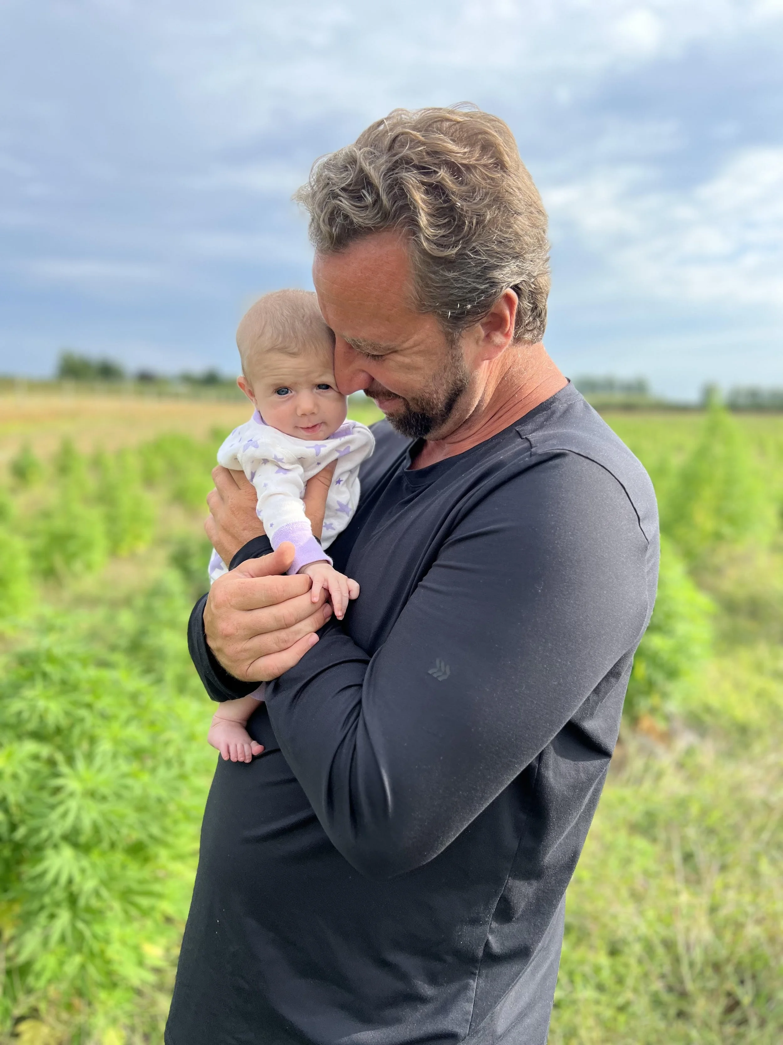 Man holding a baby outdoors in a grassy field with a cloudy sky.