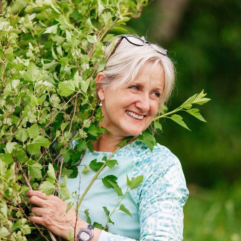 Woman smiling with short gray hair wearing a light blue shirt and holding leafy branches outdoors.