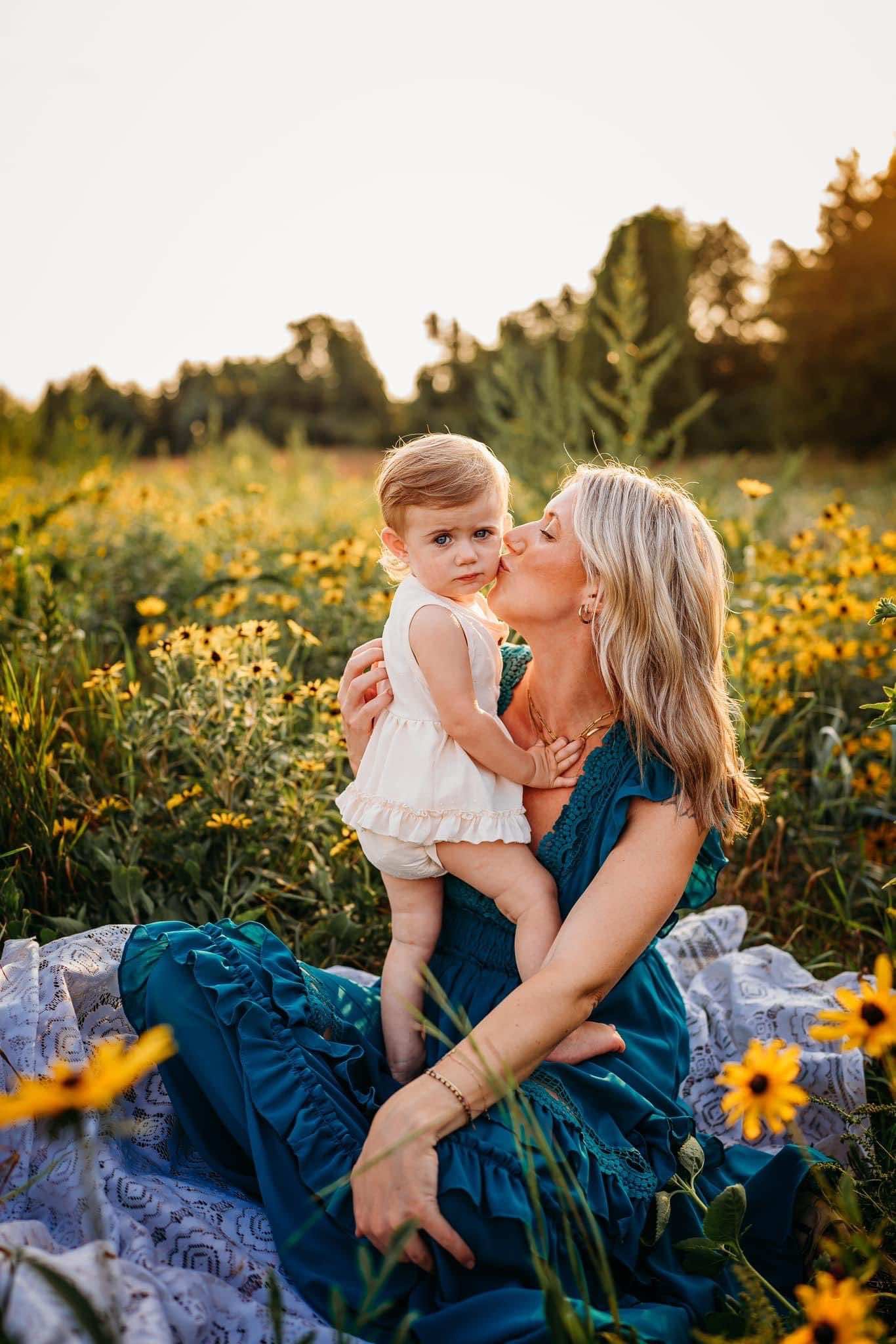 Woman in a blue dress holding and kissing a young child in a field of yellow flowers.