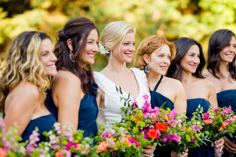 A bride in a white dress standing with bridesmaids wearing blue dresses holding colorful bouquets in an outdoor setting.