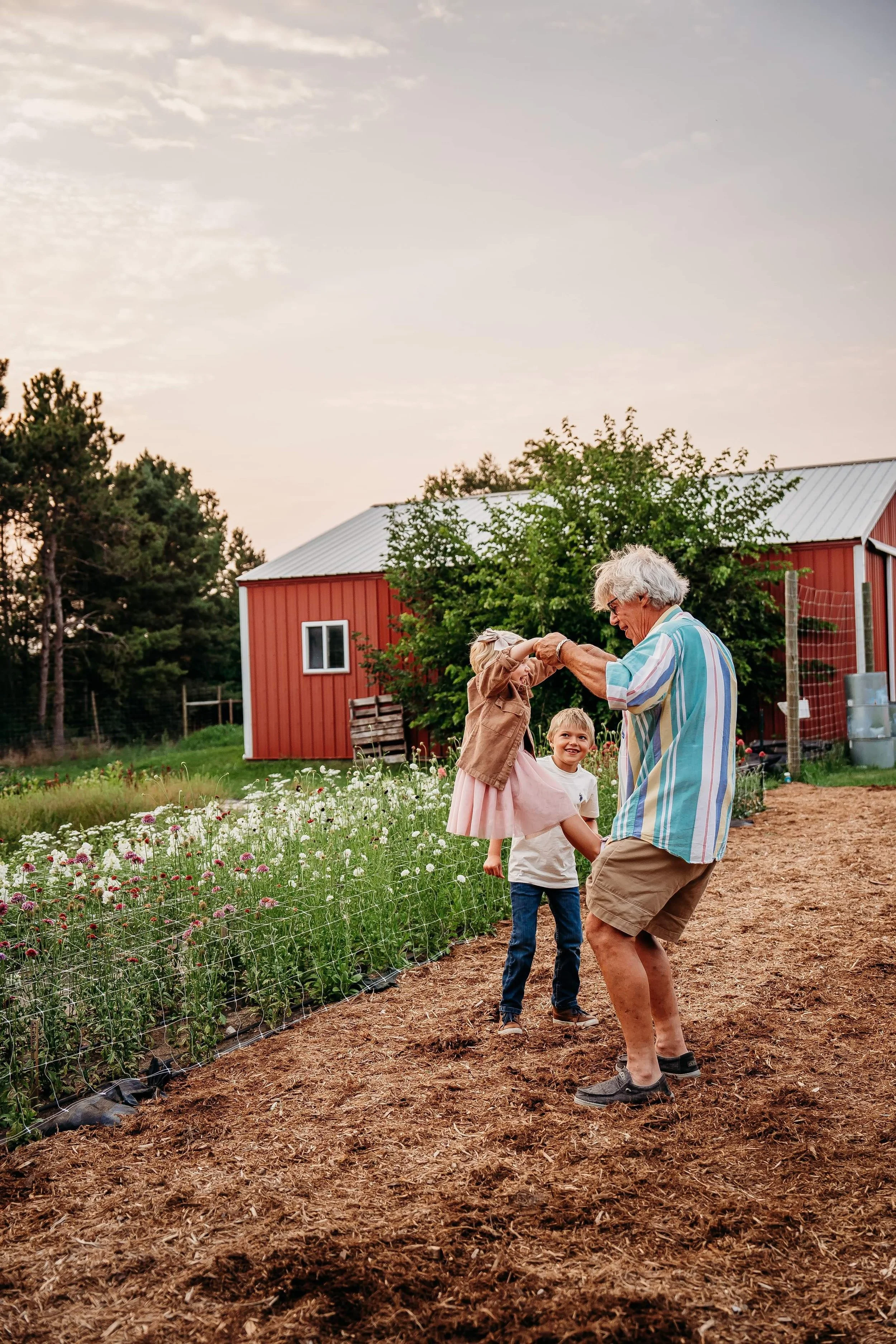A person swinging a child by the arms with another child nearby in front of a red barn and flower garden.