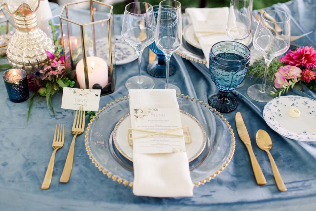 Elegant table setting with a blue tablecloth, clear charger plate, gold cutlery, and a menu card on a napkin. Crystal glasses and a blue goblet are present. A lantern with a candle, floral arrangements, and an ornate pitcher complete the setting.