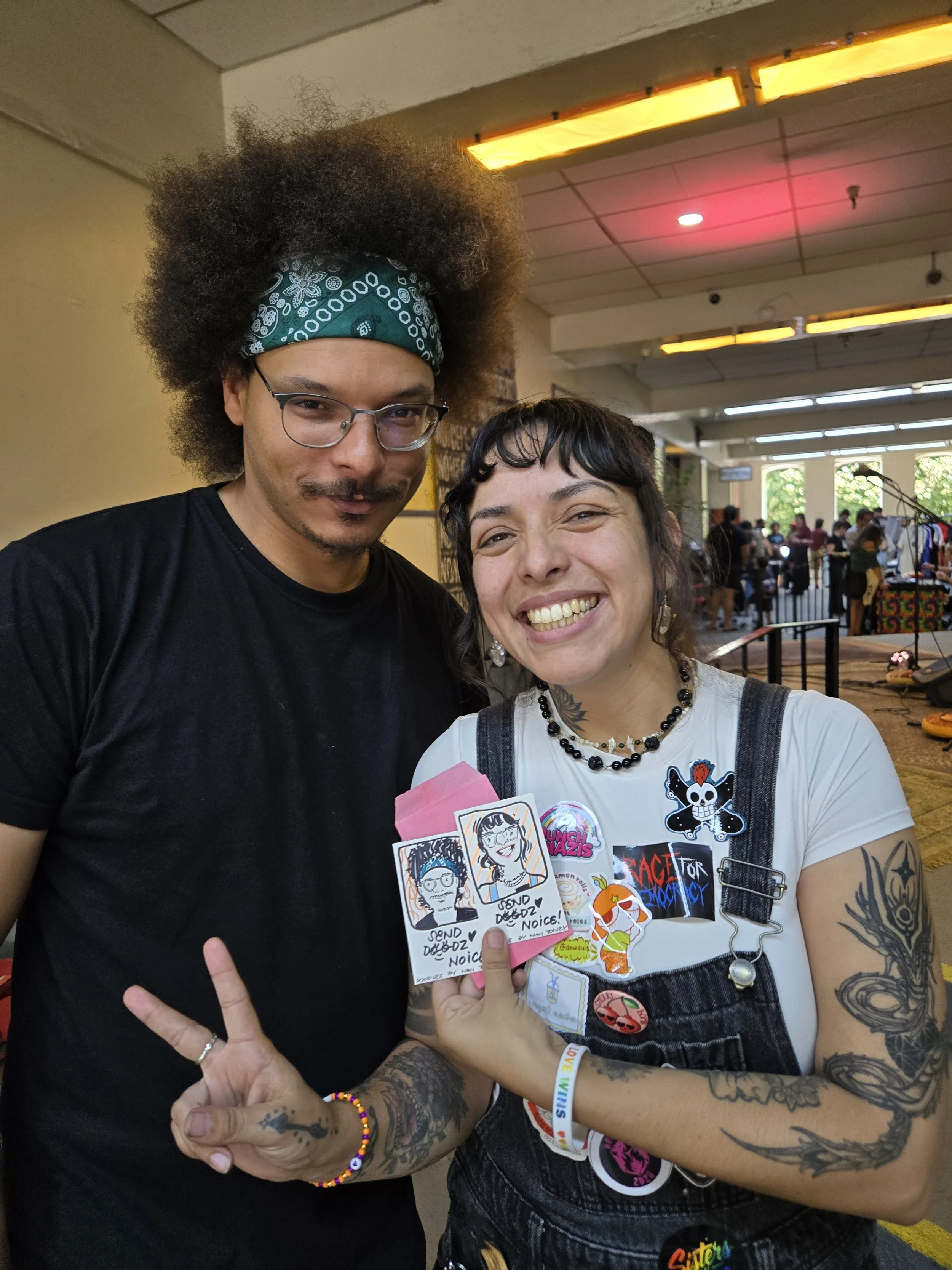 Two people smiling, one with glasses and a green bandana, the other with tattoos and pins on her overalls, holding cartoon portraits and making peace signs at an indoor event.