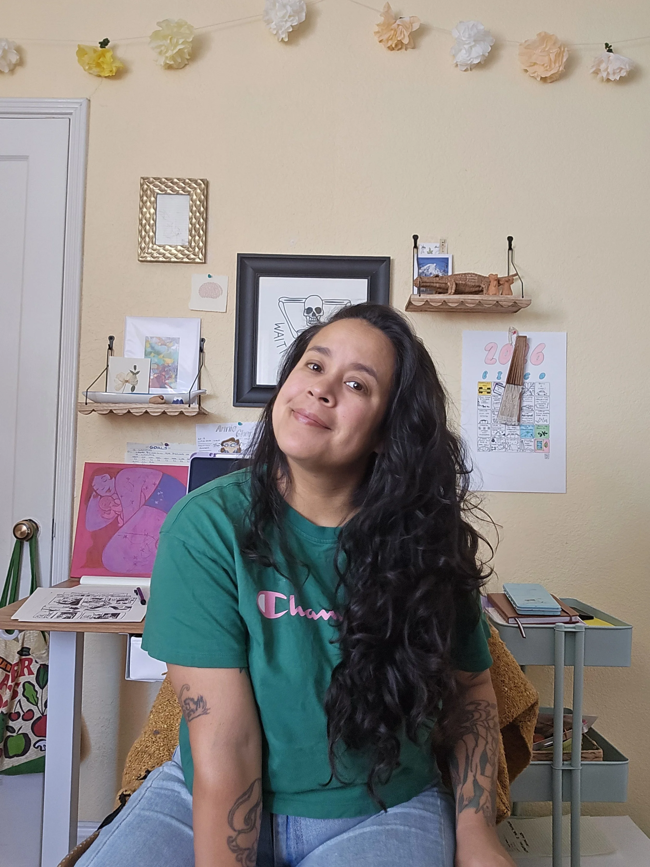 Photograph portrait of artist Niki Toney, sitting in a room decorated with hanging paper flowers and various artwork, wearing a green T-shirt and smiling at the camera.